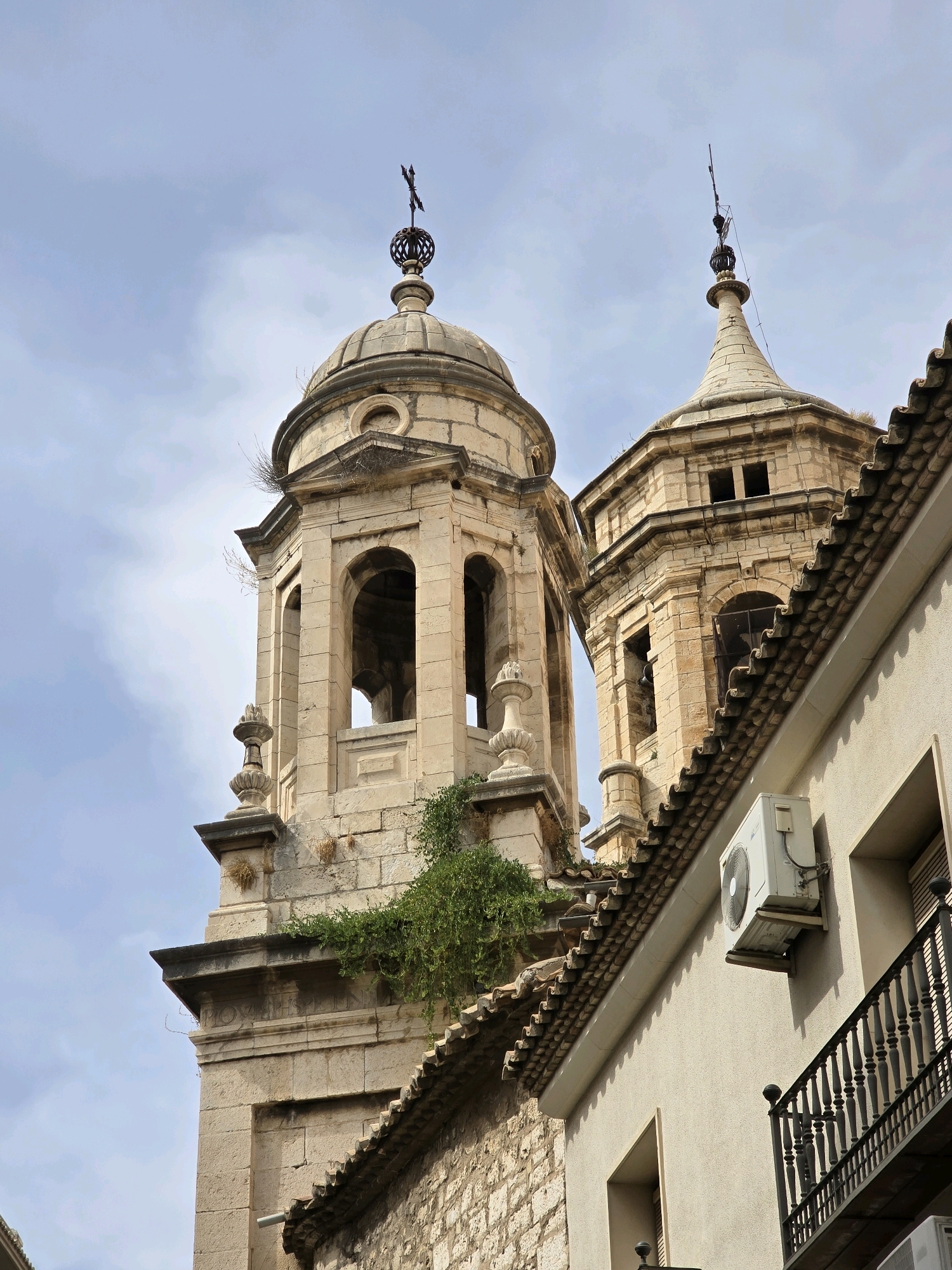 An upward, medium shot captures the upper section of two ornate, stone bell towers with domes against a pale, cloudy sky. The tower on the left is wider and appears older, with a spherical top topped by a cross. It has arched openings and decorative stone elements. The tower on the right is narrower and more conical, also topped by a cross and decorative elements. A modern, beige air conditioning unit is mounted on a light beige building in the foreground, to the right of the bell towers. The building has a dark metal railing and a tiled roof that runs diagonally across the frame. Green vines are growing on the stone of the bell towers. The overall scene depicts a juxtaposition of historic architecture and modern utility.