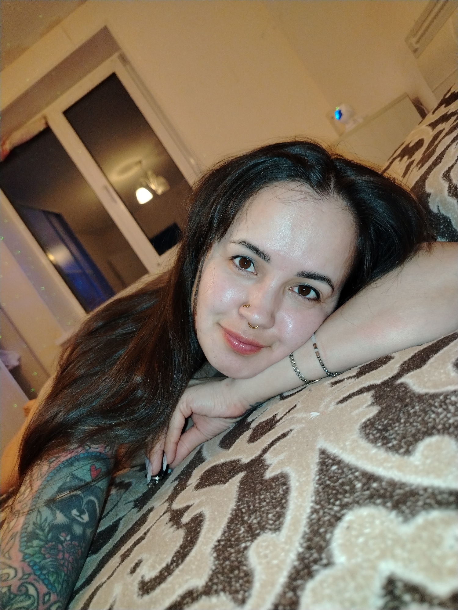A woman with long, dark hair is resting on a patterned surface, likely a couch or bed. She is fair-skinned, has brown eyes, and is wearing a nose ring. Her left arm, visible from the elbow down, features a large, colorful tattoo. She is wearing two bracelets on that wrist. A window is visible in the background. The location in Noviy, Russia cannot be determined from this image.