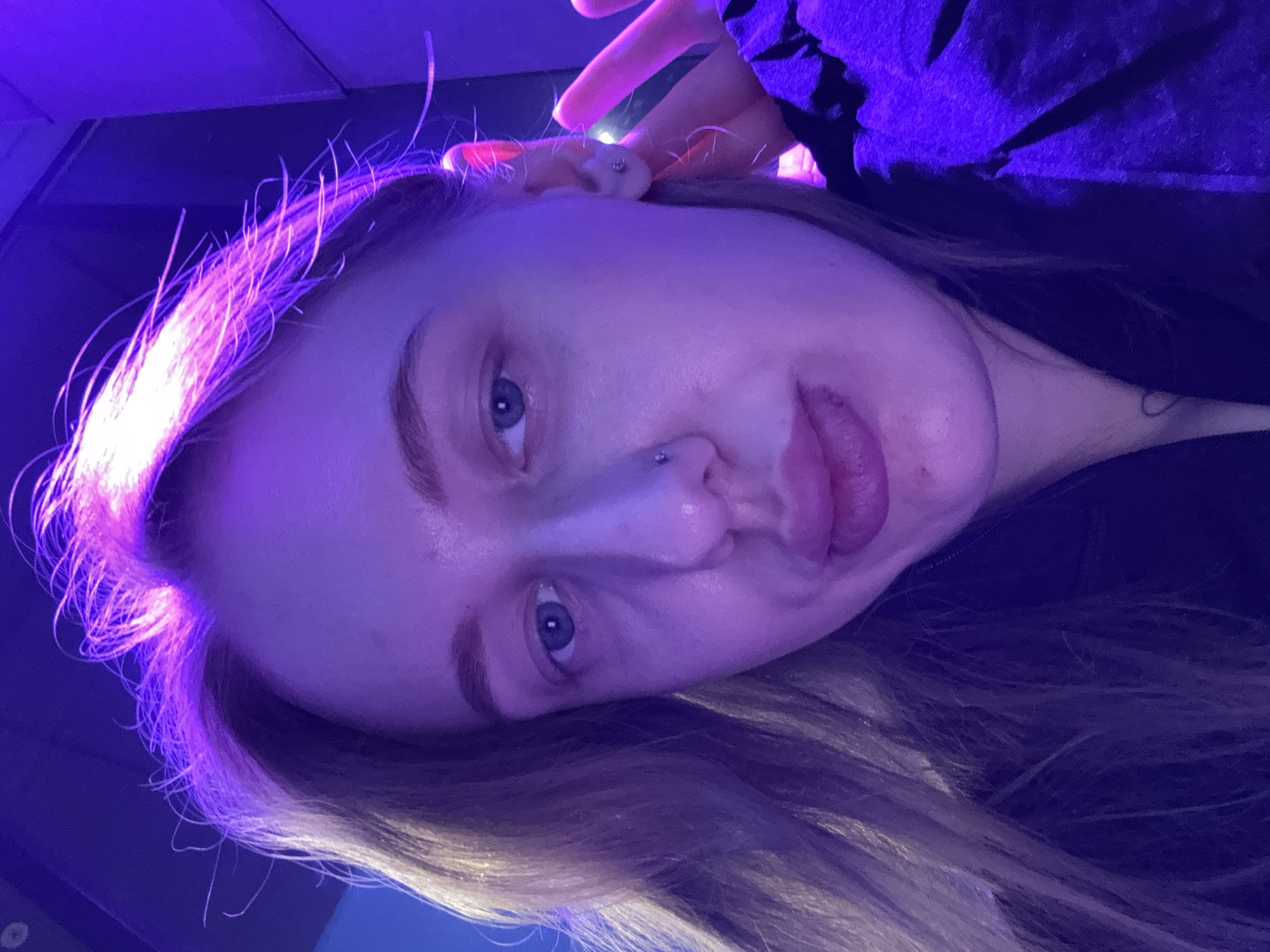 A close-up, angled photograph features a light-skinned person with blonde/brown hair, possibly female, under purple and blue lighting. The person has blue eyes and a nose piercing. Their tongue is slightly protruding from their mouth. Part of their hand is visible near their ear, with a possible piercing. The background is dark and indistinct, suggesting an indoor setting. No specific details indicate the location of Ol'ginskaya, Russia.