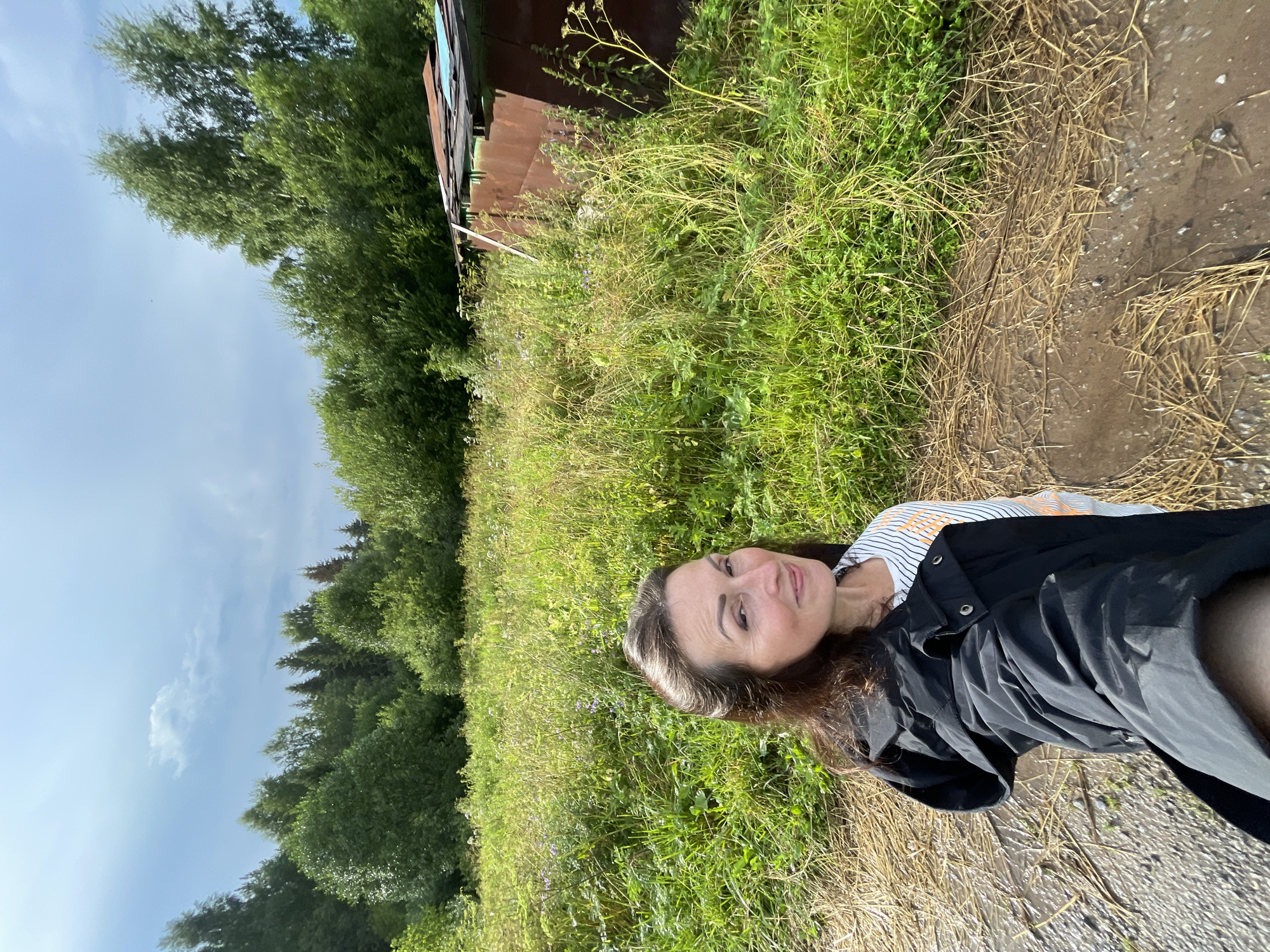 A woman is posing for a selfie in a field with trees in the background. She is wearing a black jacket over a striped shirt and has long brown hair. The field is overgrown with grass and some wildflowers, and a rusty shed is visible on the right side of the image. The sky is partly cloudy. The location appears to be rural, possibly on the outskirts of Дресвяново, Russia. The ground in the foreground is a dirt path with some dry grass.