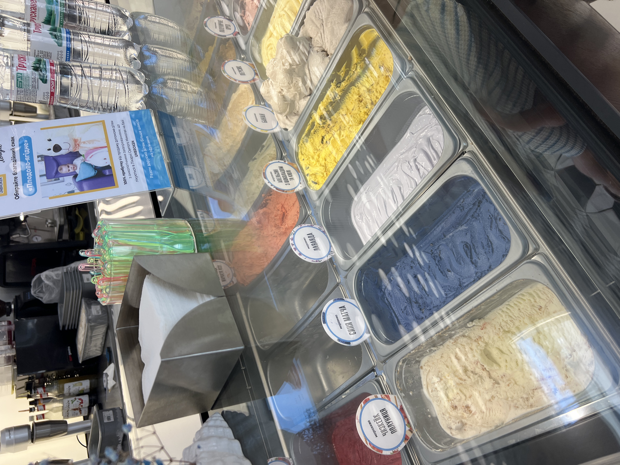 The image shows a refrigerated display case filled with various flavors of gelato or ice cream. The flavors are labeled with circular signs in Ukrainian. Some of the visible flavors include "Чизкейк Полуниця" (Cheesecake Strawberry), "Синя Мачта" (Blue Matcha), "Лаванда" (Lavender), "Апельсин з шоколадом та кюлі" (Orange with chocolate and coulis), "Банан" (Banana), and "Персик" (Peach). There are also several bottles of water with Ukrainian labels, including "Трускавецька" and "Моршинська", in the background. A promotional sign with a picture of a child and a bear, also in Ukrainian, is displayed in front of the water bottles. The environment appears to be an indoor establishment, likely an ice cream shop, with a glass counter separating the viewer from the products. The lighting is bright, reflecting off the glass and the metal of the display case.