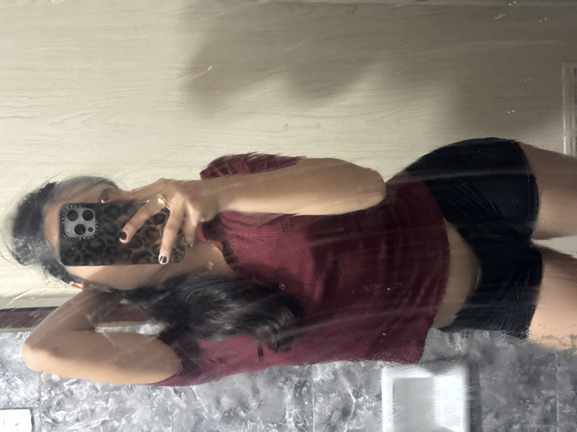 A woman is taking a mirror selfie in what appears to be a bathroom. She is wearing a short-sleeved maroon shirt and black shorts. Her hair is dark and pulled back. She is holding a phone with a leopard-print case. The background shows a marble-patterned wall and a plain wall. There is a sink visible on the left side of the image. The image is taken from the waist up. The location is not specified in the image.
