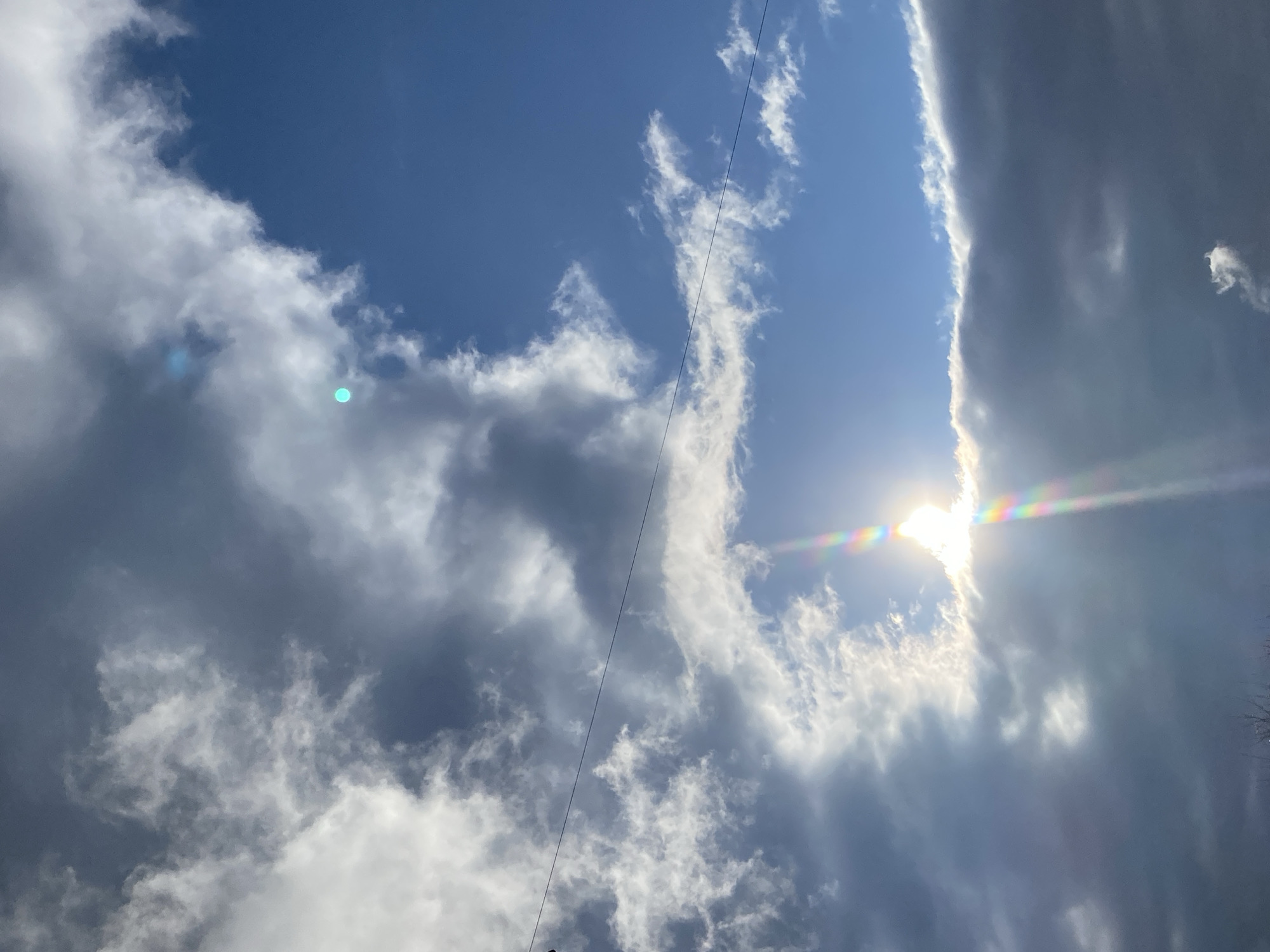 The media file displays a vibrant sky with a mix of blue sky and various cloud formations. A clear, bright blue sky is visible predominantly towards the upper left. Fluffy white clouds are scattered across the frame, intermingling with darker, denser grey clouds that dominate the lower right and center-right portions. A distinct vertical line of dark, heavy clouds stretches along the right side, illuminated by the sun's bright edge.
The sun, partially obscured by these clouds, emits a brilliant light from the lower right, producing a prominent, colorful lens flare with a rainbow spectrum that extends horizontally across the right side of the image. Several smaller, greenish-blue circular lens flares are also visible in the upper left cloud formations. A thin, dark cable, likely a utility wire, traverses diagonally across the sky from the upper central area towards the lower left. The scene, characterized by bright sunlight and varied cloud cover, depicts daytime weather conditions over Votkinsk, Russia. No visible text is present.