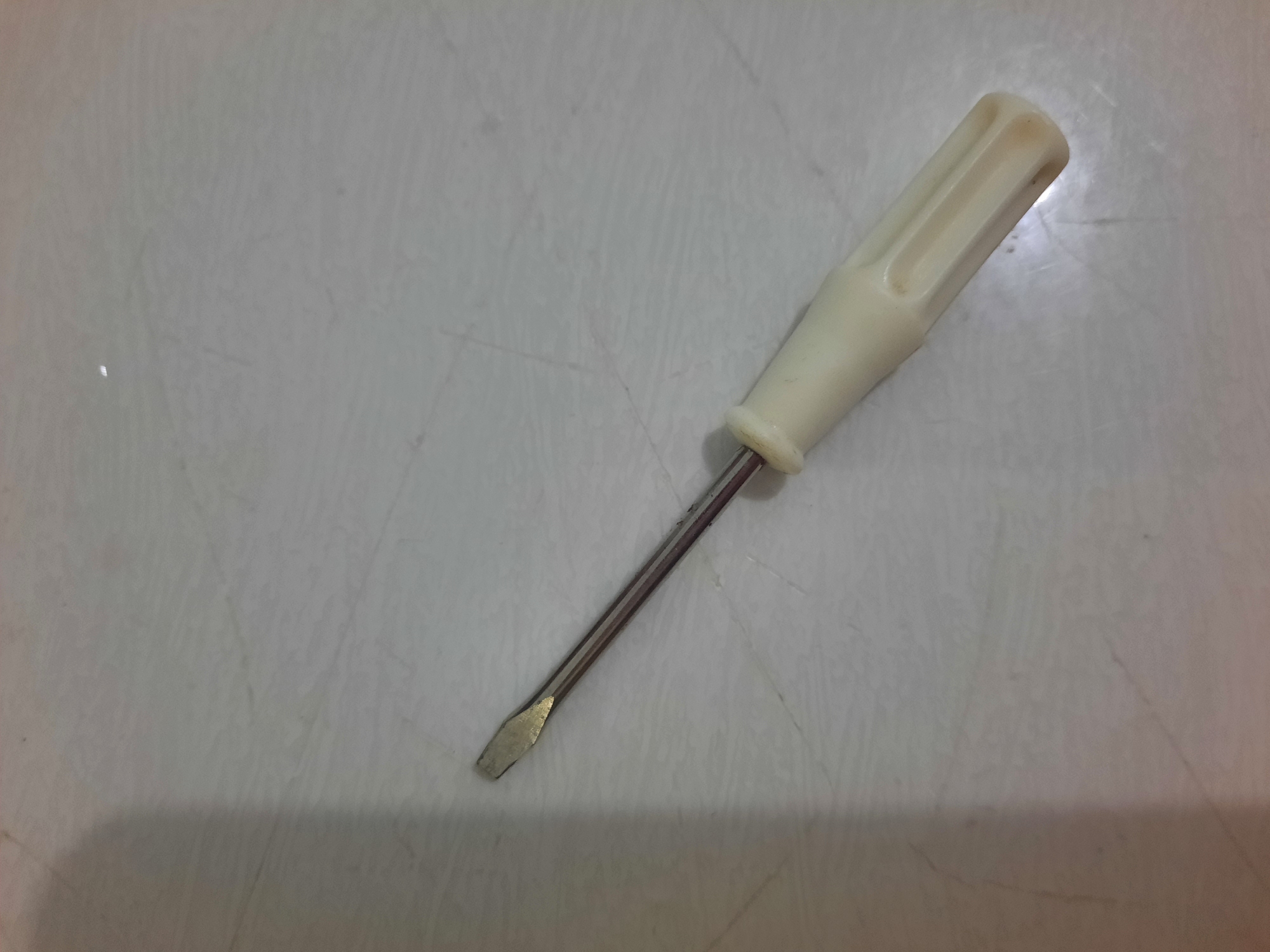 A single flathead screwdriver with a beige plastic handle lies on a light-colored, textured surface, possibly a tile floor. The screwdriver is positioned diagonally from the bottom right to the upper left of the frame. The metal shaft of the screwdriver is visible, with a shiny flathead tip. The surface it rests on has faint lines and variations in tone, suggesting it might be a countertop or flooring. There is a shadow cast on the left side of the image, indicating a light source from the upper right. No people or other objects are present. The context of Lhokseumawe, Indonesia, is not visually discernible from the image itself.