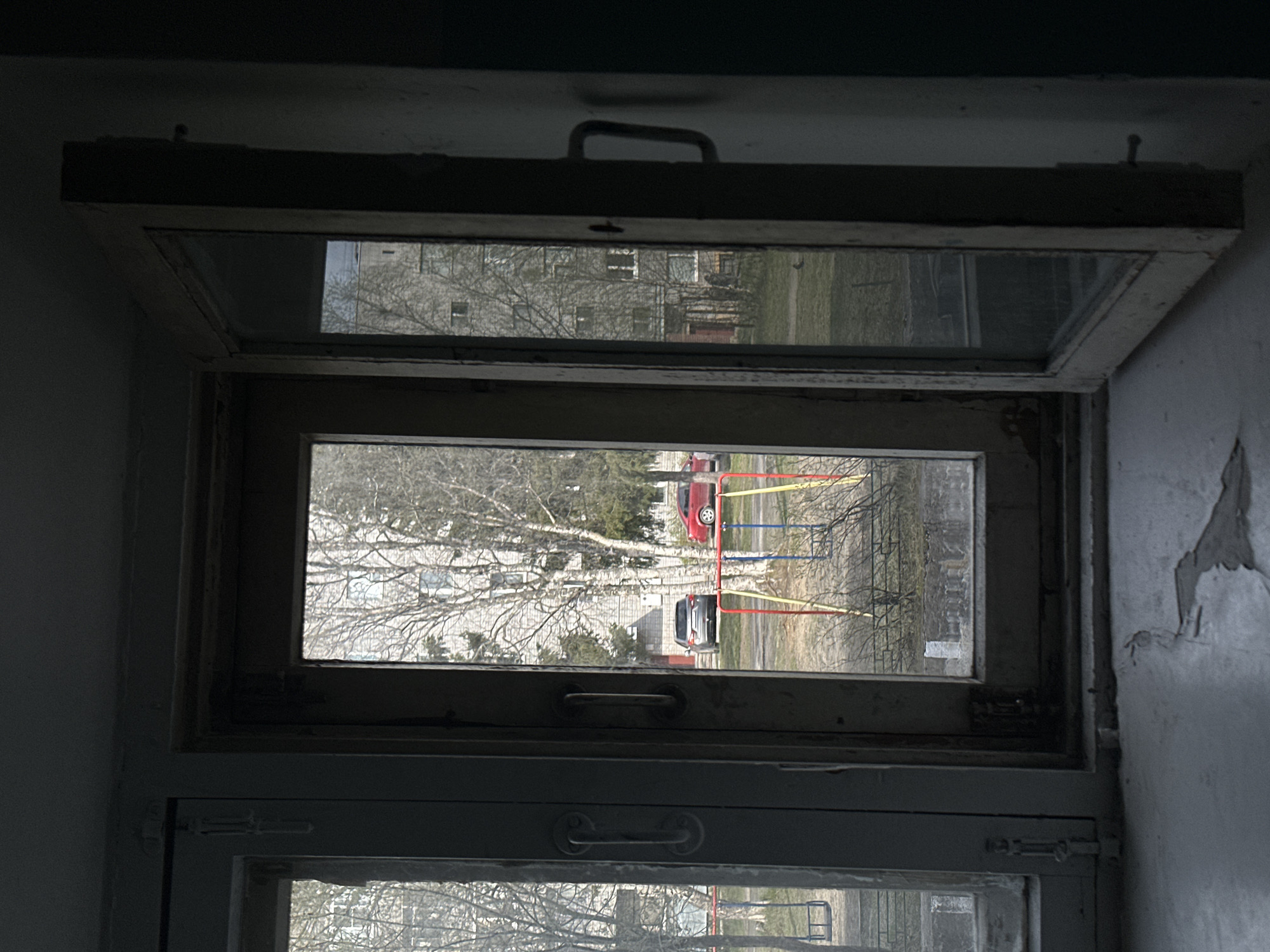 A dimly lit interior features an open, multi-paned wooden window offering a view of an urban landscape. Outside, bare trees stand in a grassy area, near a playground with brightly colored equipment. Two cars are parked on a paved surface visible through the window. The wall on the right side of the window shows signs of peeling paint and deterioration. The location is in the city of Unknown, Unknown.
