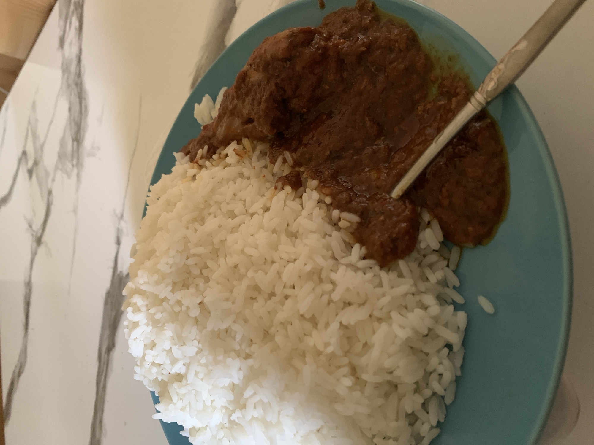 A close-up, high-angle shot shows a plate of food containing a generous portion of white rice next to a serving of brown stew. A fork rests partially submerged in the stew. The plate is teal blue and sits on a white marbled surface. The background is blurred, but appears to be a table. The scene suggests a meal in a home or restaurant setting, likely in Umuguma, Nigeria.