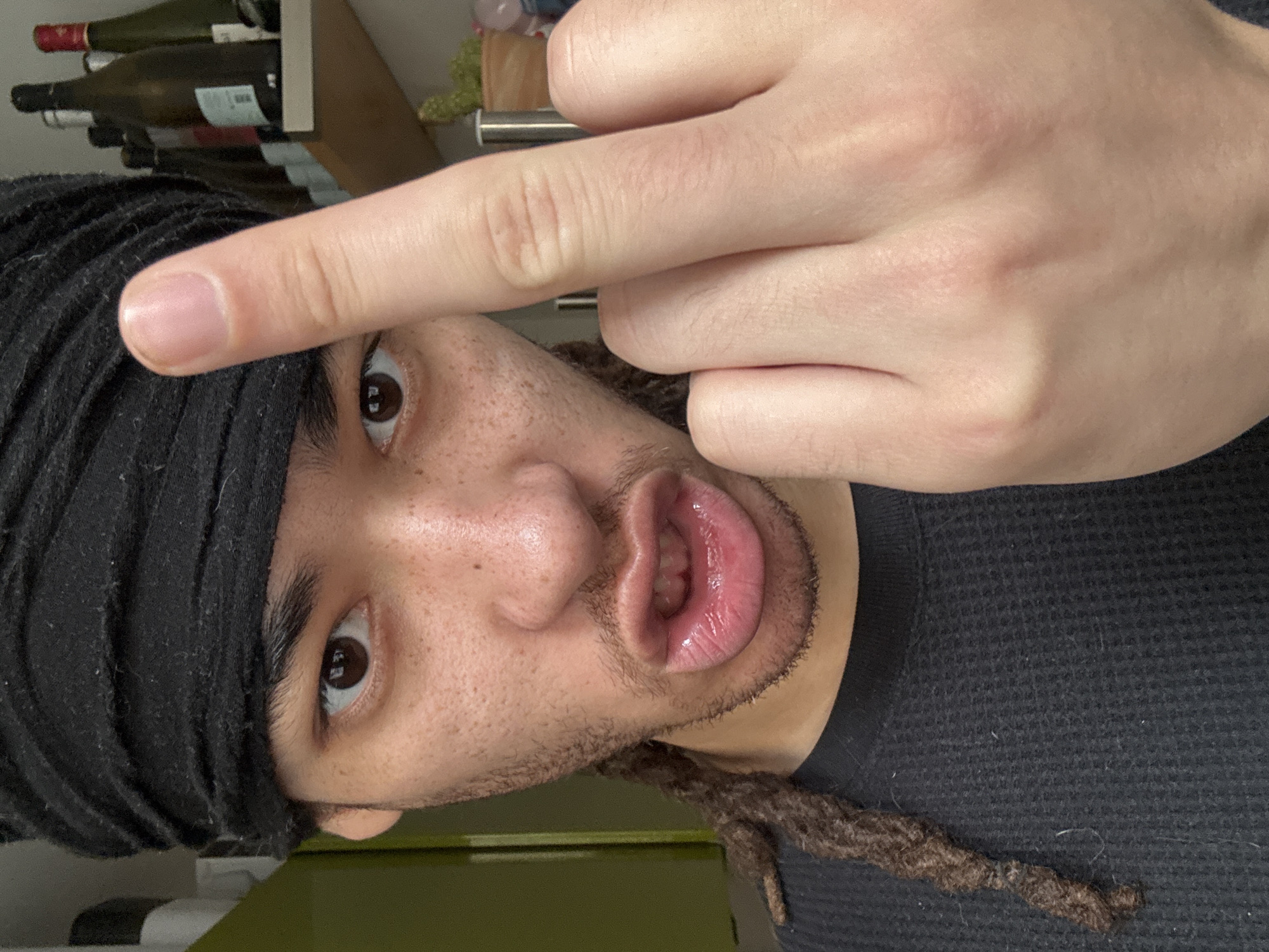 The image shows a close-up self-portrait of a person with brown eyes, freckled skin, and dreadlocks, wearing a black headwrap and a dark gray waffle-knit shirt. The person is giving the middle finger to the camera. The background is out of focus, but includes a shelf with bottles, possibly wine bottles. There is no indication of the image being in Paris, France.