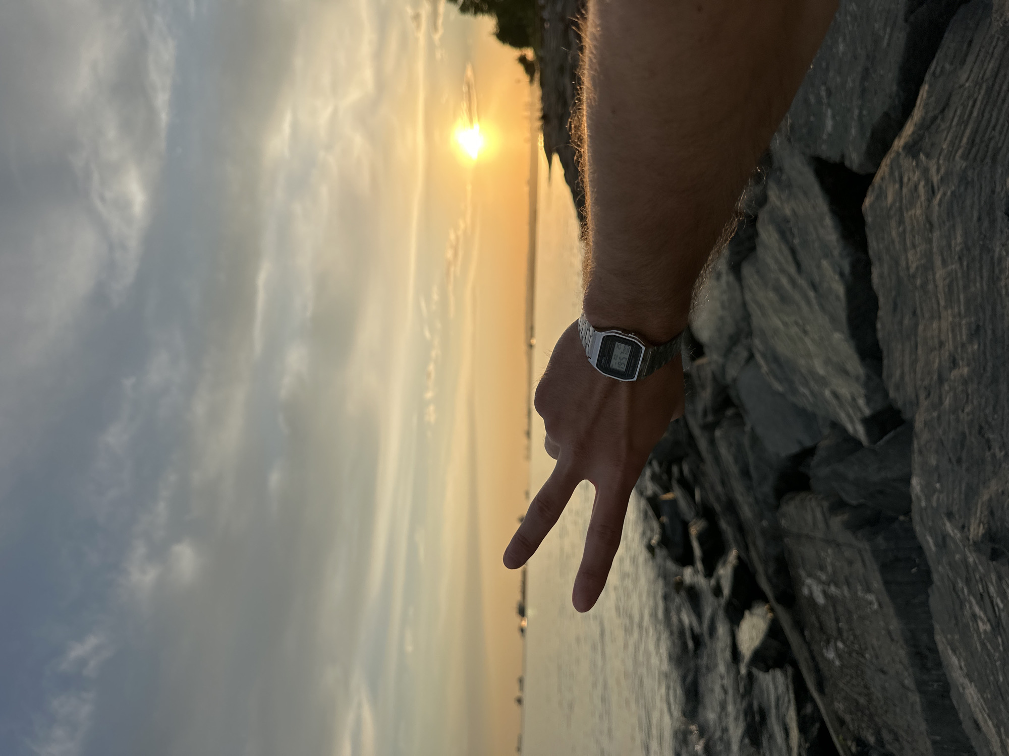 A person's arm and hand are in the foreground, with two fingers extended in a peace sign. The person is wearing a silver digital watch on their wrist. The background features a body of water, likely the Sea of Marmara, with several ships visible in the distance. A rocky shoreline is present in the lower portion of the image, and the sky is filled with clouds, with the sun setting and casting an orange glow. The scene appears to be located along the coast of Bakırköy, Turkey, given the presence of the sea and ships.