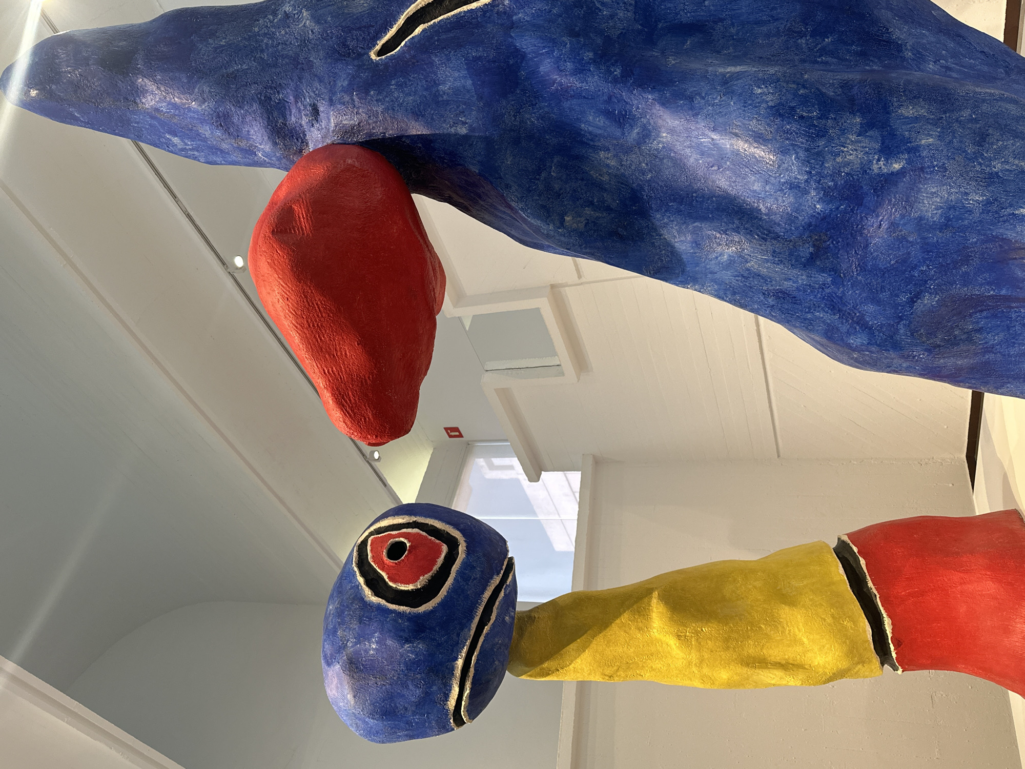 The image shows a close-up, upward-angled view of a sculpture by Joan Miró, specifically "Barcelona, 1968," located in the Fundació Joan Miró in Barcelona, Spain.
The sculpture features a combination of primary colors – blue, red, and yellow – and has a whimsical, abstract form. The blue element is large and dominant, with a rough, textured surface. A red, bulbous shape appears to be connected to or hanging from the blue form. Lower in the frame is a blue rounded head with a distinctive eye marked by concentric rings of red, black, and white. A yellow arm or appendage extends from the head, transitioning into red at the end. The sculpture is set against a backdrop of a bright, white interior, possibly the museum's ceiling or a skylight, with architectural details visible. The orientation of the sculpture is unusual, likely suspended from the ceiling.