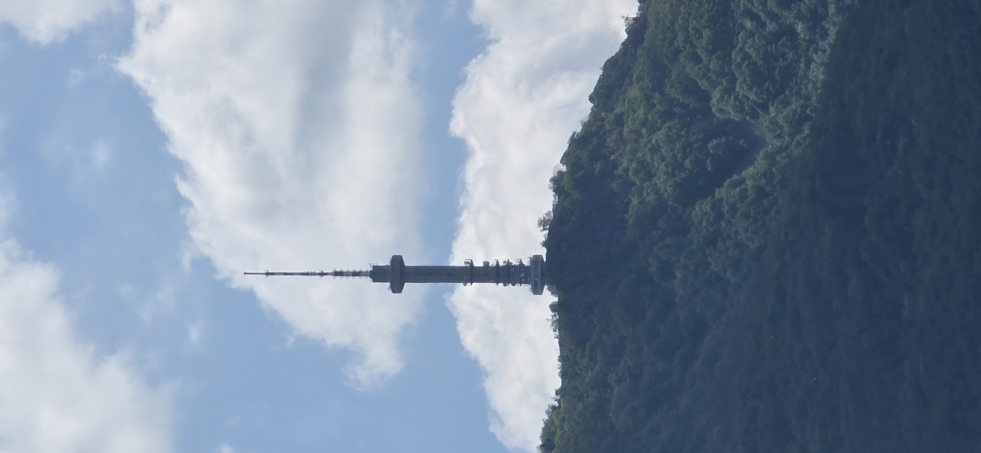 The image depicts a telecommunications tower located on Mount Vitosha, near Sofia, Bulgaria. The tower is situated on the ridgeline of the mountain, appearing as if it connects the mountain to the sky filled with clouds. The mountain is heavily forested with green trees.