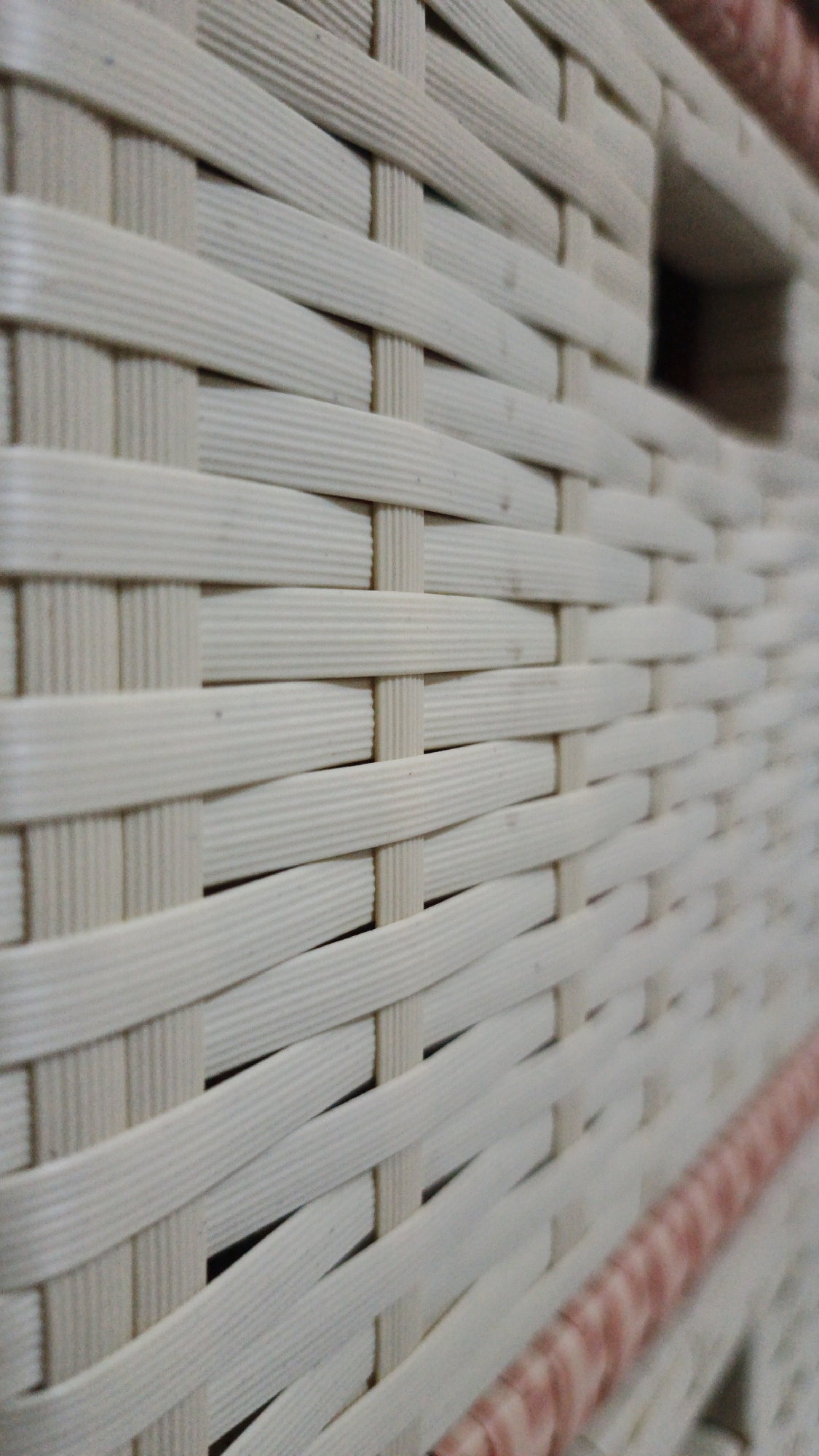 The image is a close-up, angled shot of a woven basket. The basket is made of light-colored, possibly plastic or resin, material that is woven in a tight criss-cross pattern. The weave is textured with fine parallel lines running along the length of each strand. In the upper right portion of the image, there is a rectangular opening or hole in the basket's weave. Along the bottom right edge of the image, a decorative band of pink and white striped material is visible, suggesting the basket might be for decoration or storage. The background is mostly out of focus, showing the continuation of the woven pattern and hints of darker space within the basket. There are no people or animals present. The image does not contain any discernible landmarks or context that would place it specifically in Muntur, Indonesia. It is a generic close-up of a woven object.