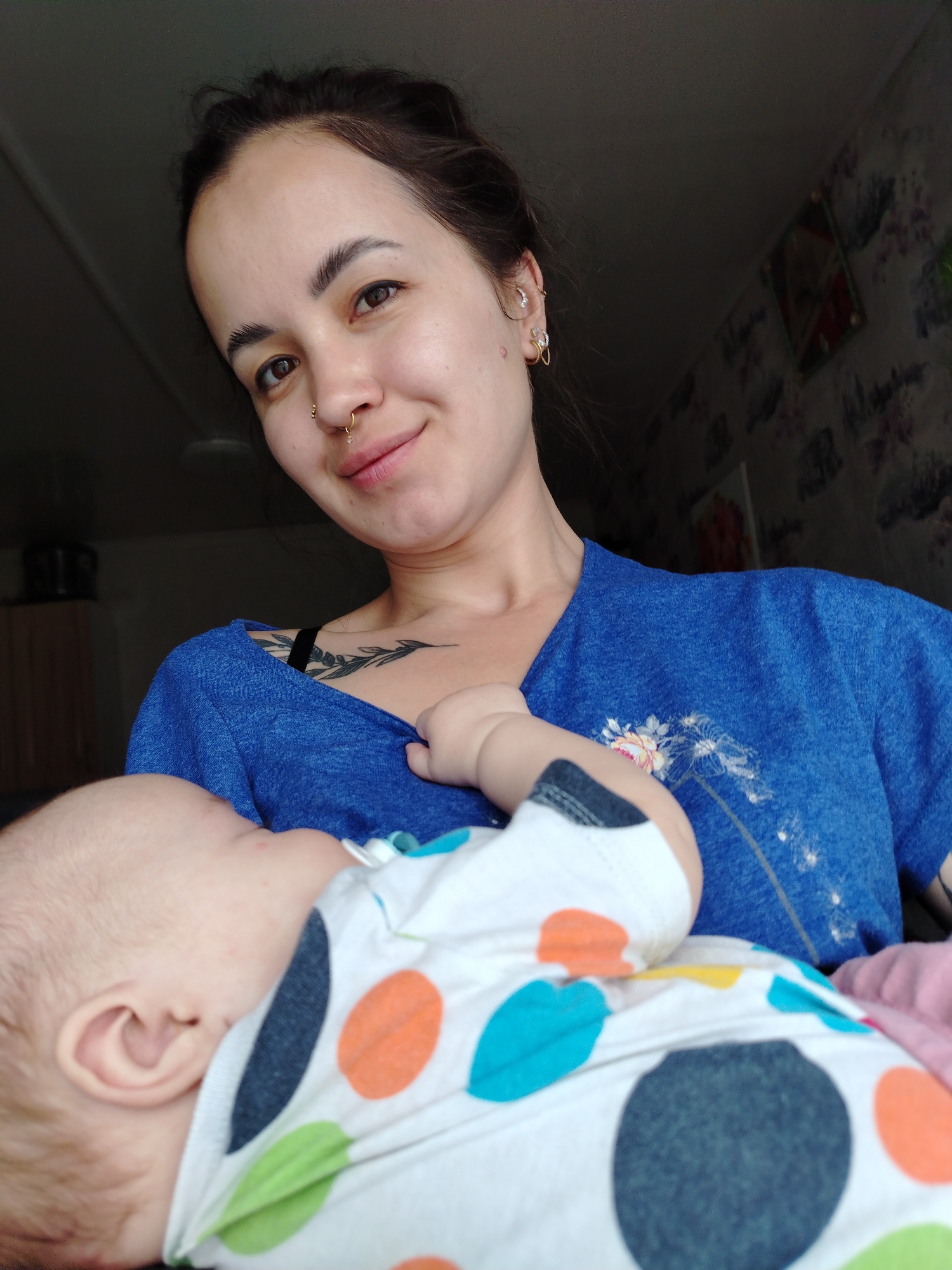 The photograph depicts a woman holding a baby. The woman has dark hair pulled back, light skin, and pierced nostrils and ears. She is wearing a blue T-shirt with a floral design. A tattoo is visible on her upper chest. The baby is dressed in a white outfit with colorful polka dots and a pink blanket. The location appears to be indoors, with wallpaper and furniture visible in the background. There is no verifiable information on the location being in Гожан, Russia. The woman is looking at the camera and appears to be smiling. The baby is facing away from the camera.