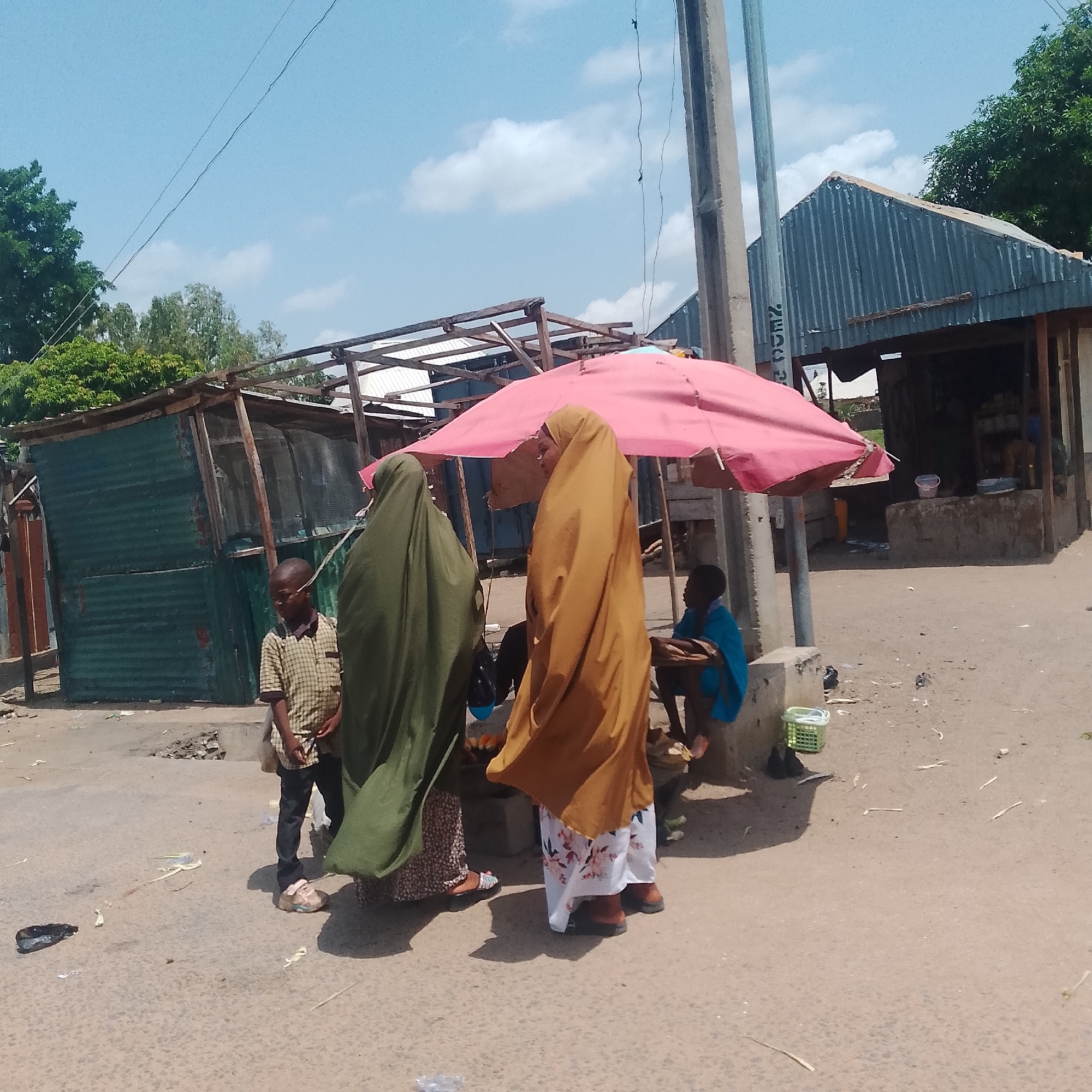 A street scene in Jalingo, Nigeria, features two women in traditional Islamic attire, one in green and one in a golden-brown hijab, standing near a market stall. A young boy, wearing a checkered shirt and dark pants, stands to their left, facing away from the camera. To the right, a child in a blue garment sits under a large pink umbrella, which also shelters the market stall. The stall has a rough, concrete counter and appears to be selling items, with buckets visible. Behind the stall, a corrugated metal building forms the background, with a wooden structure and trees visible beyond. The ground is paved and shows signs of debris. A utility pole stands to the right of the scene. The sky is blue with scattered clouds, indicating daylight. The overall impression is a busy, open-air market setting.