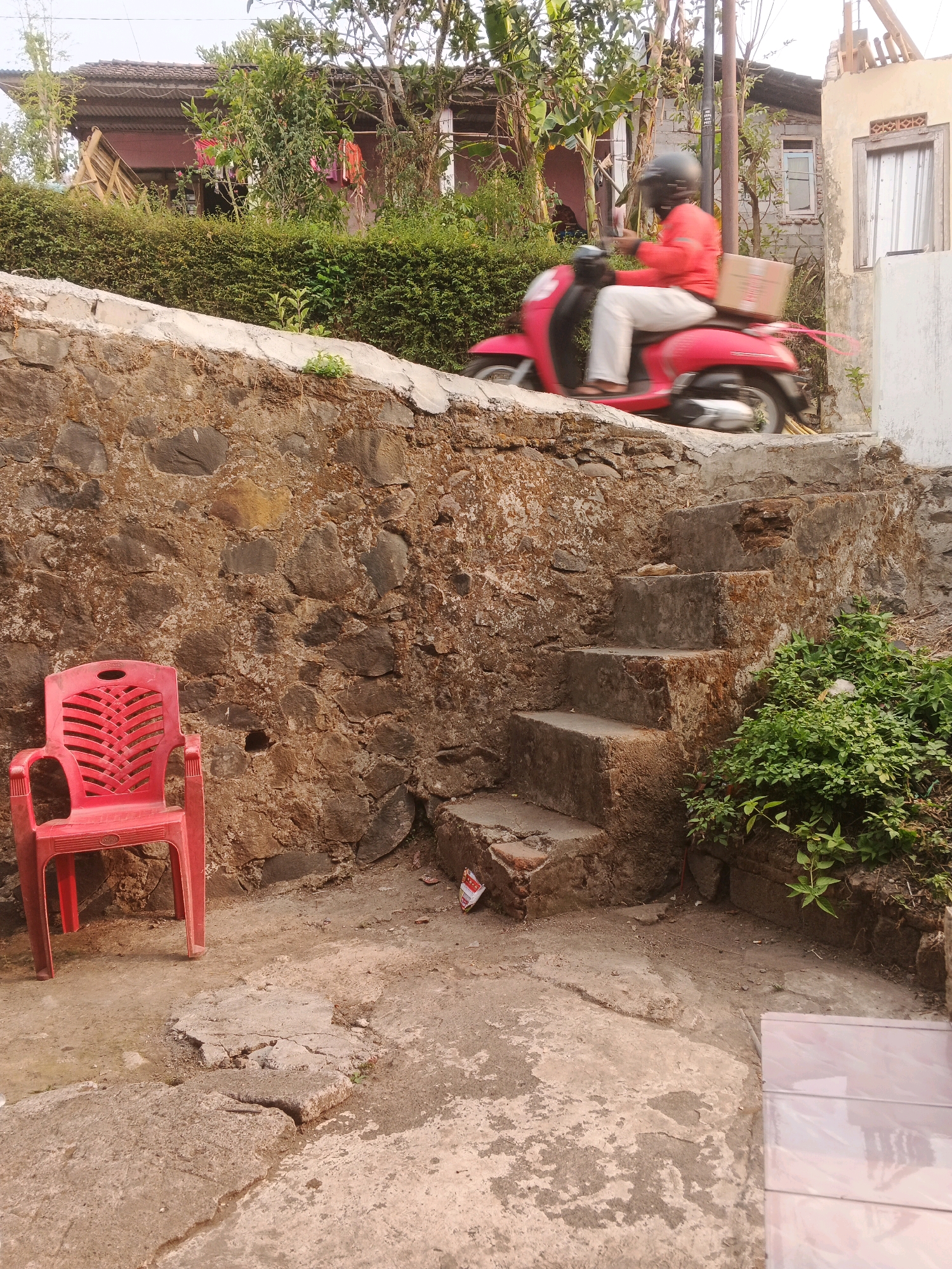 A person is riding a red scooter with a box on the back, moving from left to right on a paved surface. They are wearing a helmet and an orange jacket. The scooter is slightly blurred due to motion. In the foreground, to the left, is a red plastic chair placed on a concrete ground. Behind the chair, a textured stone wall rises. To the right of the stone wall, there is a set of concrete stairs leading upwards. Overgrown green bushes are visible to the right of the stairs. The background shows a building with a corrugated roof and vegetation. The scene appears to be in a residential area, possibly in the city of Keji, Indonesia, given the architectural style and lush greenery.