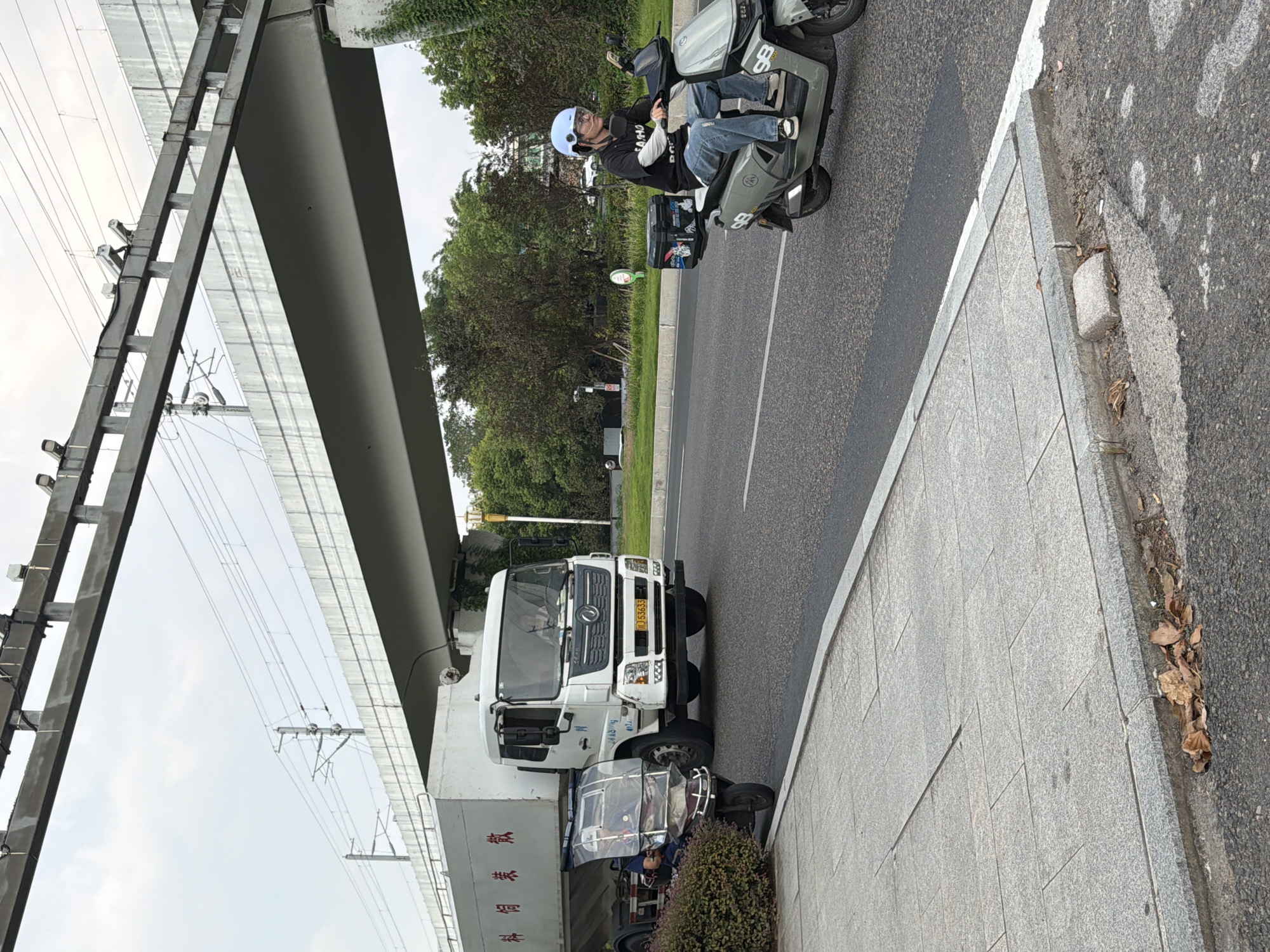 The image depicts an outdoor scene in the city of Jiangjin, China, under an overpass, likely for a road or railway. In the foreground, a person wearing a blue helmet, glasses, a black t-shirt, and blue jeans is riding a light gray electric scooter. The scooter has a black storage box mounted on the rear. The person is looking forward and appears to be in motion on the road. To the left, a white truck with a large white cargo container is parked or moving slowly. The cargo container has red Chinese characters on its side. Next to the truck, a three-wheeled electric scooter with a clear windshield is visible, with a person sitting on it. The area to the left of the road features a sidewalk with gray paving stones and some fallen leaves near the curb. Trees and vegetation are visible behind the road and under the overpass. The sky is overcast.