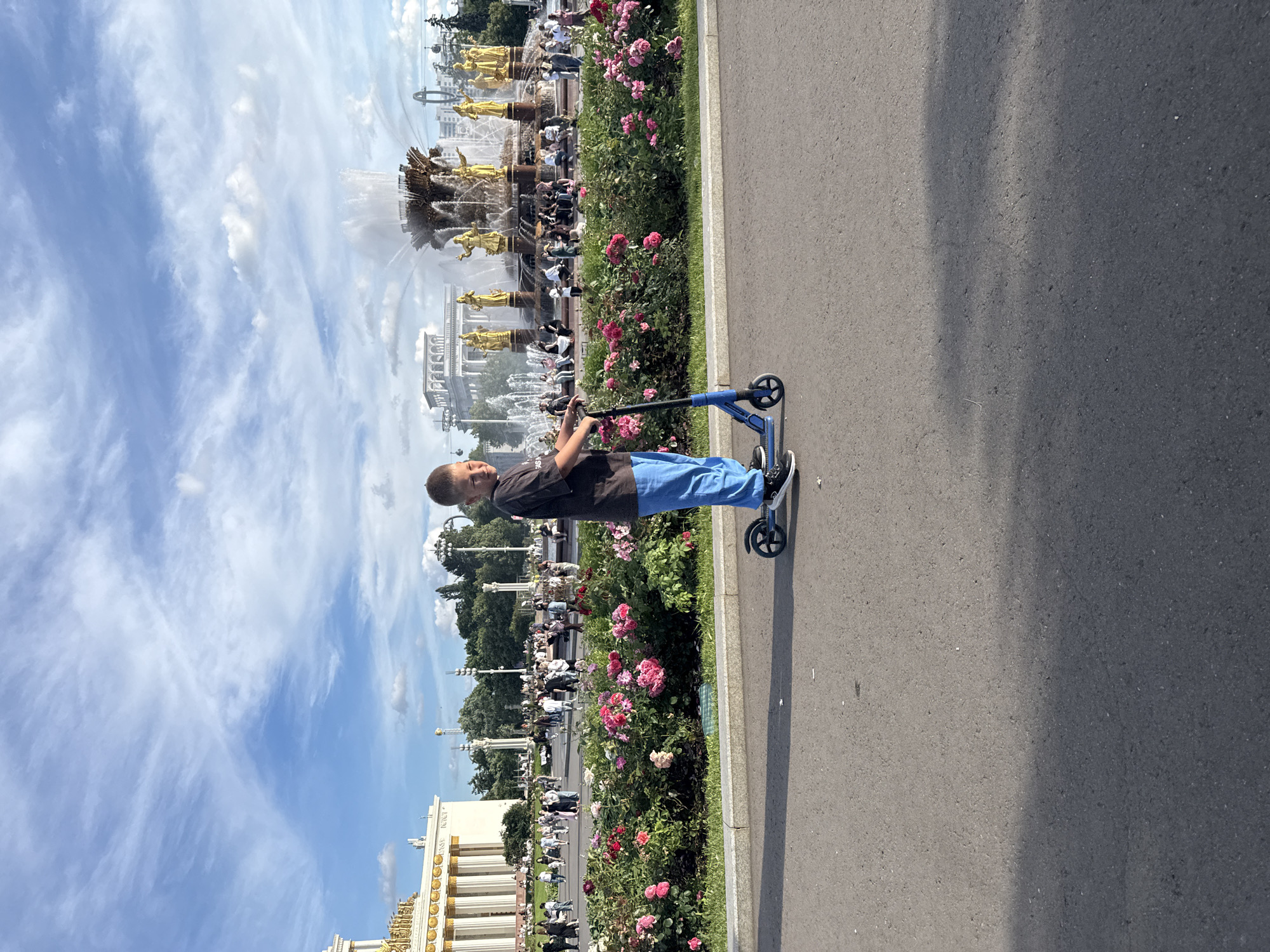 A young boy rides a blue scooter on an asphalt pathway in VDNKh park in Moscow, Russia. He is wearing a dark t-shirt and blue pants. In the background, a large fountain with golden statues is visible, along with ornate buildings and a crowd of people enjoying a sunny day. A flower bed with pink roses is in front of the fountain. The sky is blue with wispy white clouds.