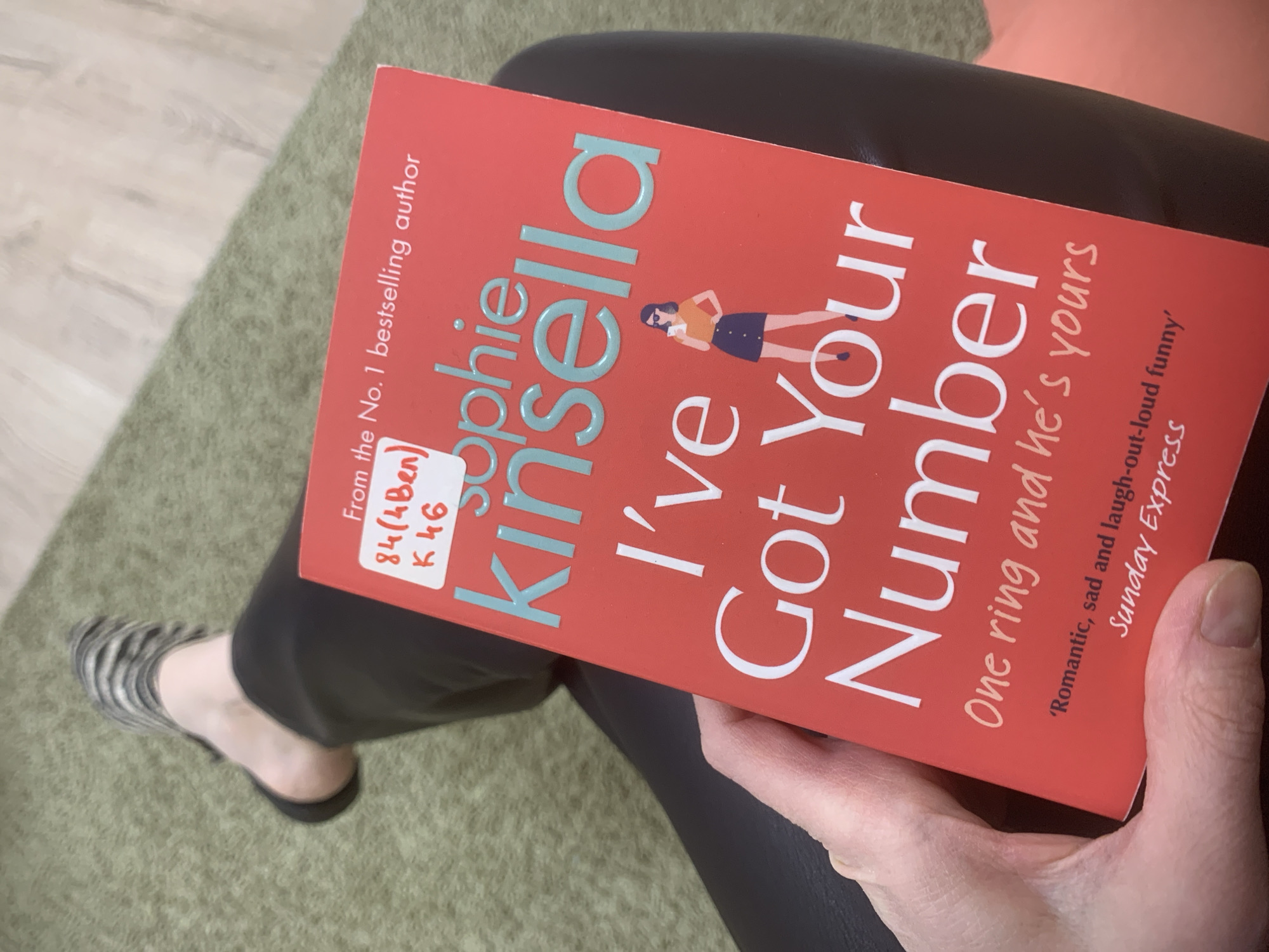A woman is sitting down, holding a book titled "I've Got Your Number" by Sophie Kinsella. The book has a red cover with the title in white and light blue font. She is wearing black pants. Her left foot is visible, clad in a zebra-striped sandal. The background includes a green rug and a partially visible orange cushion. The book has a library sticker. There is no indication of Izhevsk, Russia.