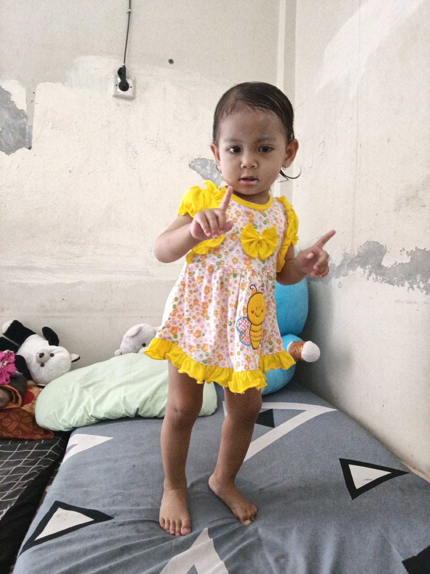 A young child, appearing to be around two years old, stands on a bed in a room. The child is wearing a yellow and white floral dress with a yellow bow and a bumblebee applique. The child's hair is dark and slicked back, and they are looking directly at the camera with a neutral expression. Their right arm is raised with the index finger pointing upwards, and their left arm is extended with the index finger pointing outwards.
The bed has a grey and white patterned comforter. To the left of the child, on the bed, are several stuffed animals, including a panda and a white bear. A light green pillow is also visible.
The wall behind the child is off-white and appears to be textured with some peeling paint or plaster in places. An electrical outlet with a black cord running from it is mounted on the wall above and to the left of the child. To the right of the child, a large blue stuffed toy is partially visible.
The scene appears to be an indoor residential setting, likely a bedroom. Based on the visual cues and the user's prompt, this scene is located in Denpasar, Indonesia.