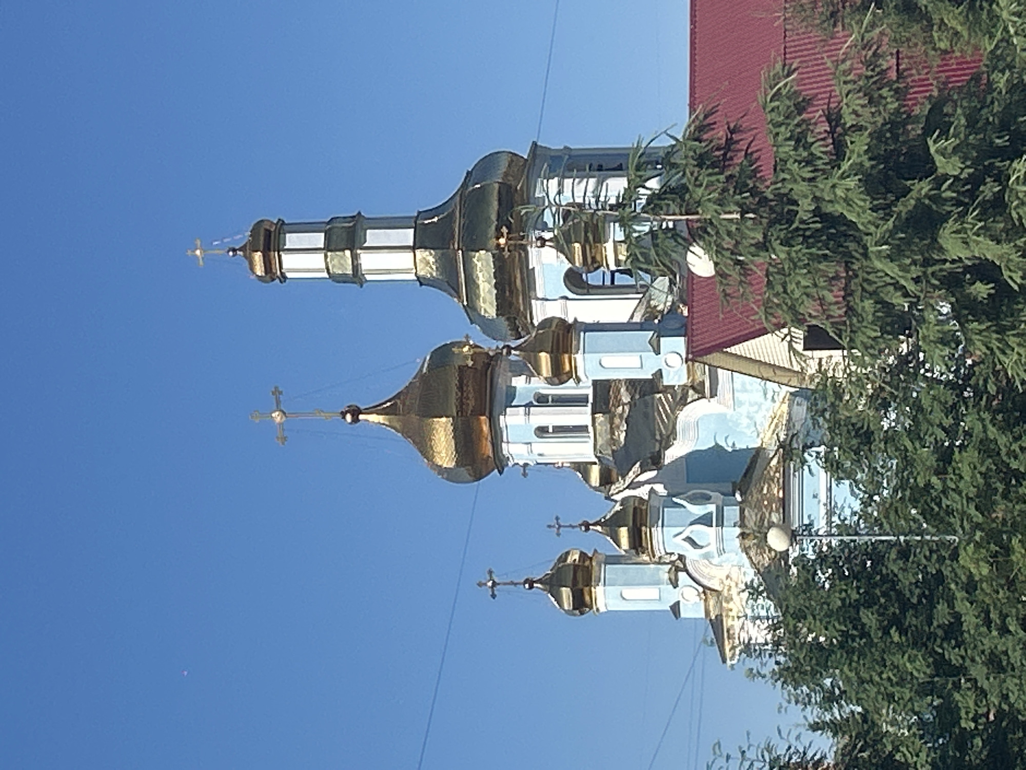 This is a vertical, medium shot photograph of the Ascension Cathedral in Kemerovo, Russia, against a clear blue sky. The cathedral features multiple golden domes and crosses, and its lower sections are painted light blue and white. A tall, slender tower also topped with a golden dome and cross rises on the right side of the building. In the foreground, lush green trees partially obscure the lower part of the cathedral. To the right, a section of a building with a red roof is visible. The overall scene depicts a sunny day with clear visibility.