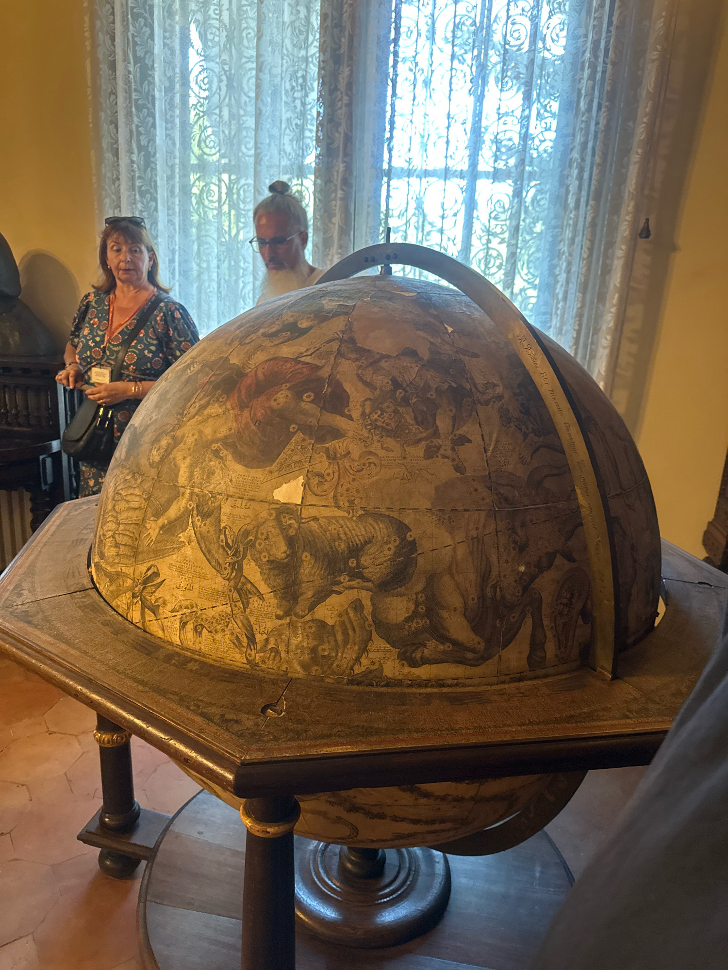 The image shows a large, antique celestial globe displayed on a wooden stand. The globe is decorated with intricate illustrations of constellations and mythological figures. Two individuals are partially visible in the background, standing near a window with sheer curtains. A woman in a patterned dress is on the left, and a man with a bun and beard is on the right. The scene appears to be indoors, in a room with tiled floors and muted lighting. The location is not identifiable as Belgrade, Serbia.
