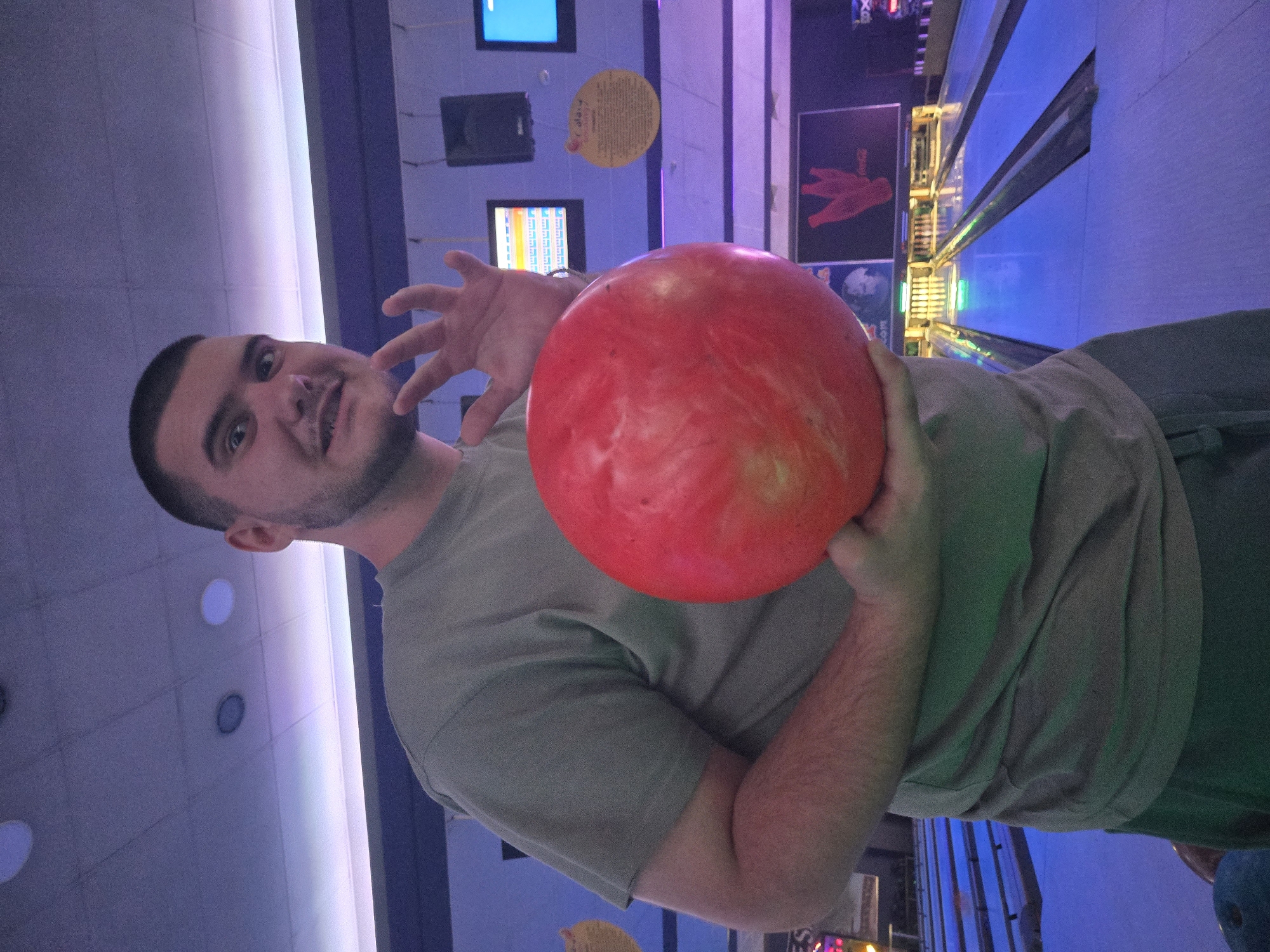 A man holds a red bowling ball in a bowling alley. He has dark hair and a beard. He is wearing a green t-shirt and grey pants. The background shows bowling lanes, monitors, and colorful lighting. The bowling alley is located in Pleven, Bulgaria.