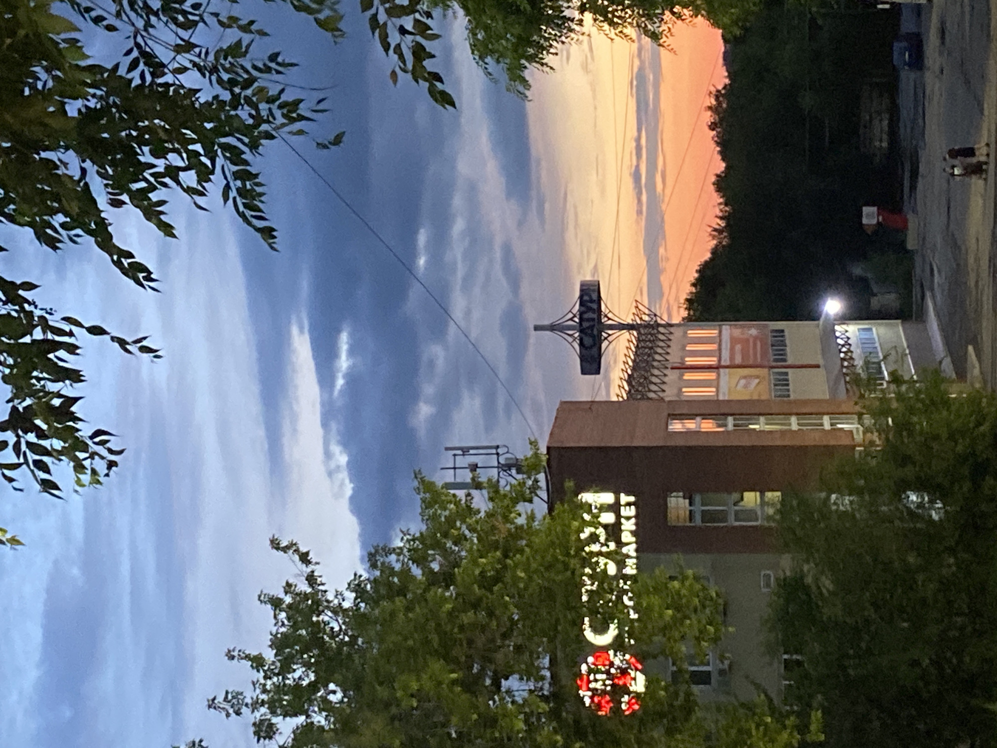 This is a vertical, medium-distance shot of a street scene in Novokuybyshevsk, Russia, captured at dusk. The sky displays a dramatic blend of colors, with dark blue clouds transitioning to a warm orange and yellow sunset near the horizon.
In the foreground, green foliage from trees frames the left and bottom portions of the image, partially obscuring the view of a building. The building, a multi-story structure, appears to be a commercial establishment. A brightly lit sign on its facade reads "СПАР МАРКЕТ" (SPAR MARKET), with the word "СПАР" (SPAR) in a white, stylized font and "МАРКЕТ" (MARKET) in a vertical white font. Below "СПАР", there's a circular red illuminated logo. Another sign with horizontal orange and white stripes is visible higher up on the building.
A tall metal structure with antennas is also visible above the trees and next to the building. In the background, a darker line of trees separates the buildings from the vibrant sky. Further to the right, a paved area resembling a street or parking lot is visible, with two indistinct figures walking away from the camera. A street lamp is illuminated on the right side of the image. The overall mood is peaceful, with the warm sunset colors contrasting with the darkening sky and the cool blue tones. The time of day is clearly evening, as indicated by the sunset and the illuminated signs.