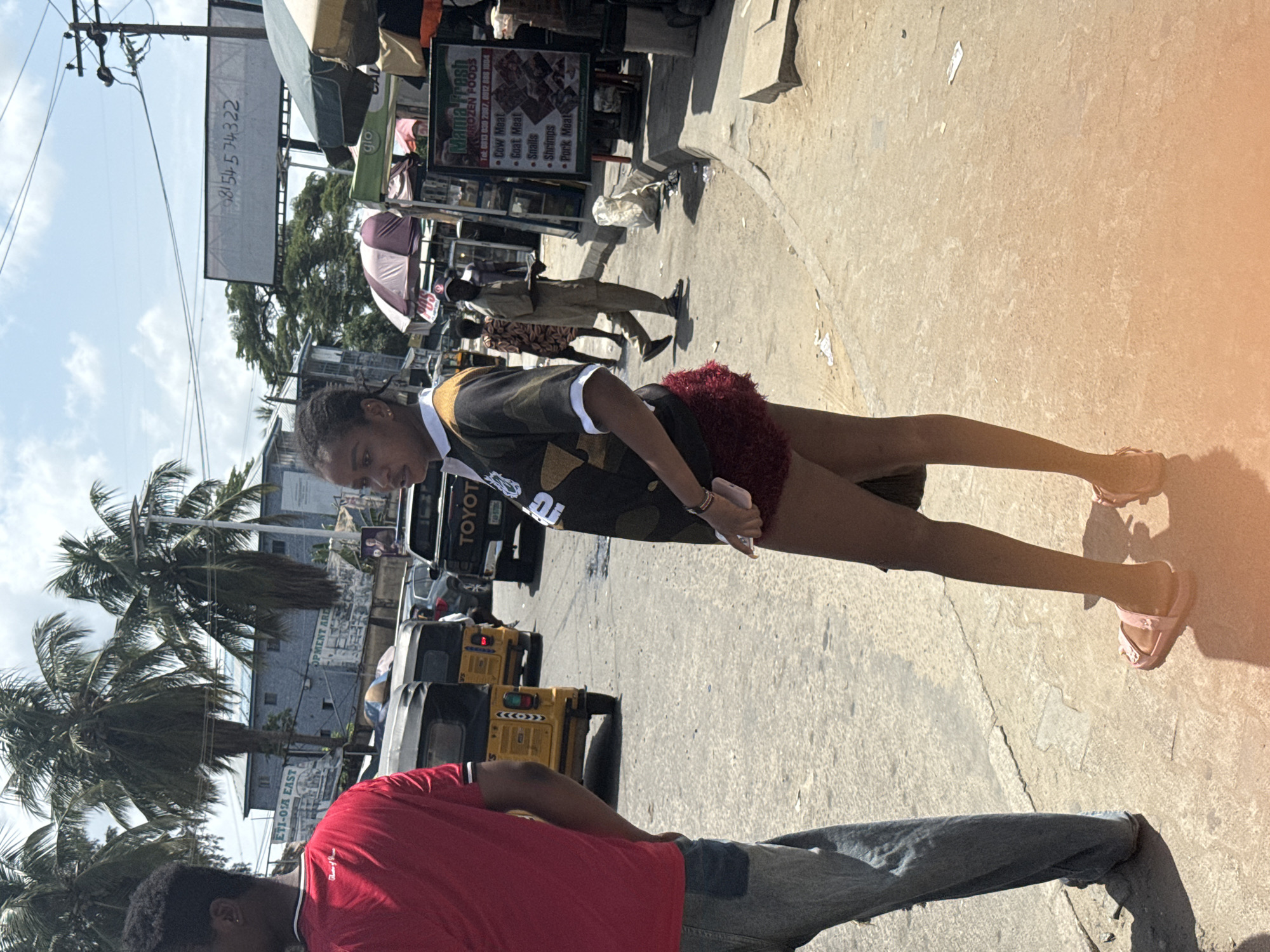 The image captures a street scene in Ado, Nigeria. A woman in a black and camouflage shirt and burgundy shorts stands with a phone in her hand. Beside her is a man in a red shirt and jeans. A Toyota car and yellow tricycles pass behind them. Palm trees are visible in the background. Street vendors and pedestrians are seen near stalls with signs advertising Mama Fresh Frozen Foods. Utility poles and overhead wires are prominent.