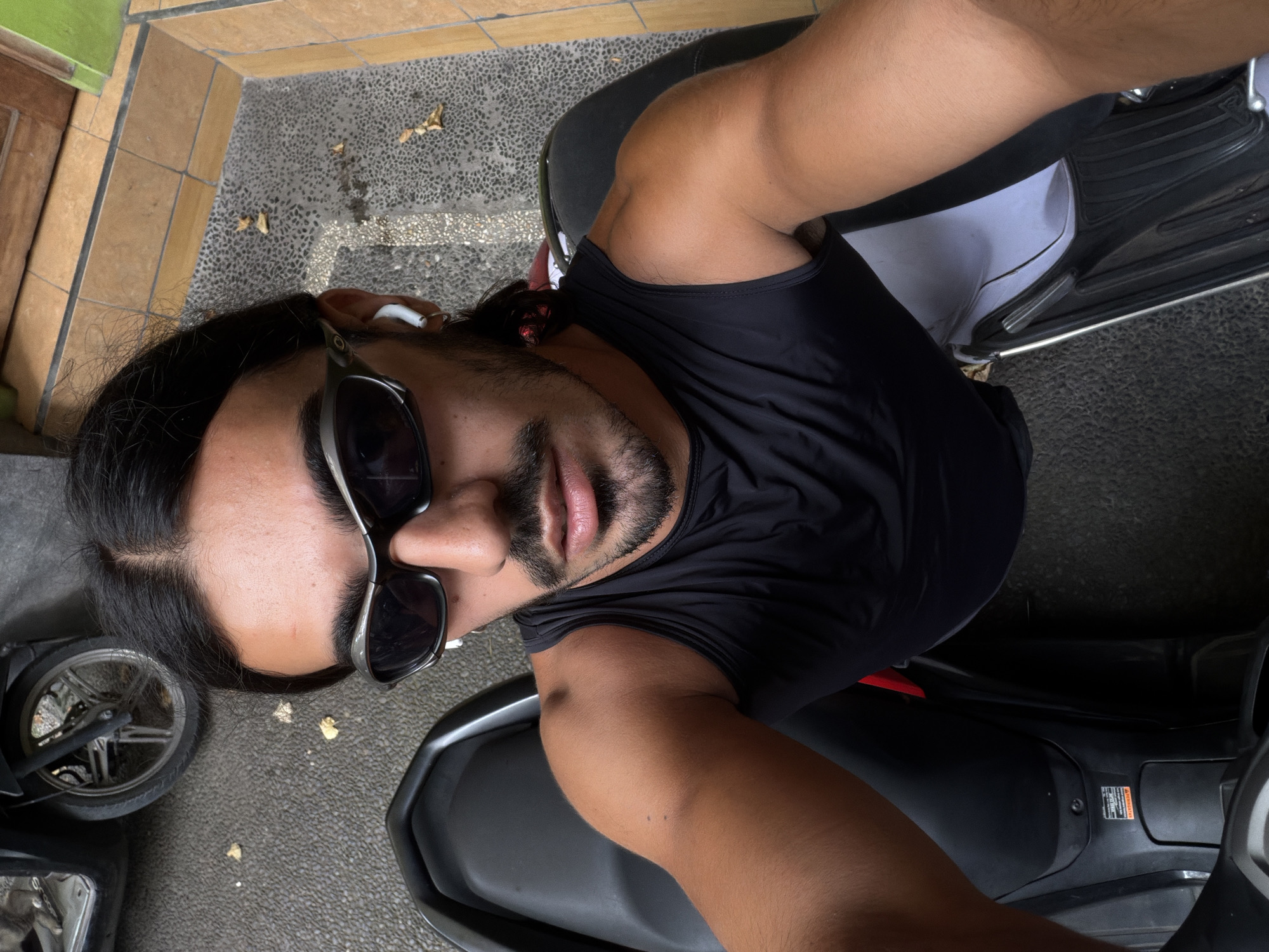 A man wearing sunglasses and a black tank top is seated on a parked motorcycle in North Kuta, Indonesia. He appears to be taking a selfie from an overhead angle. His skin tone is warm, and he has a beard and long hair. He is wearing a white earbud in his left ear. Part of a building's exterior and a section of a paved area can be seen in the background, along with another parked motorcycle.