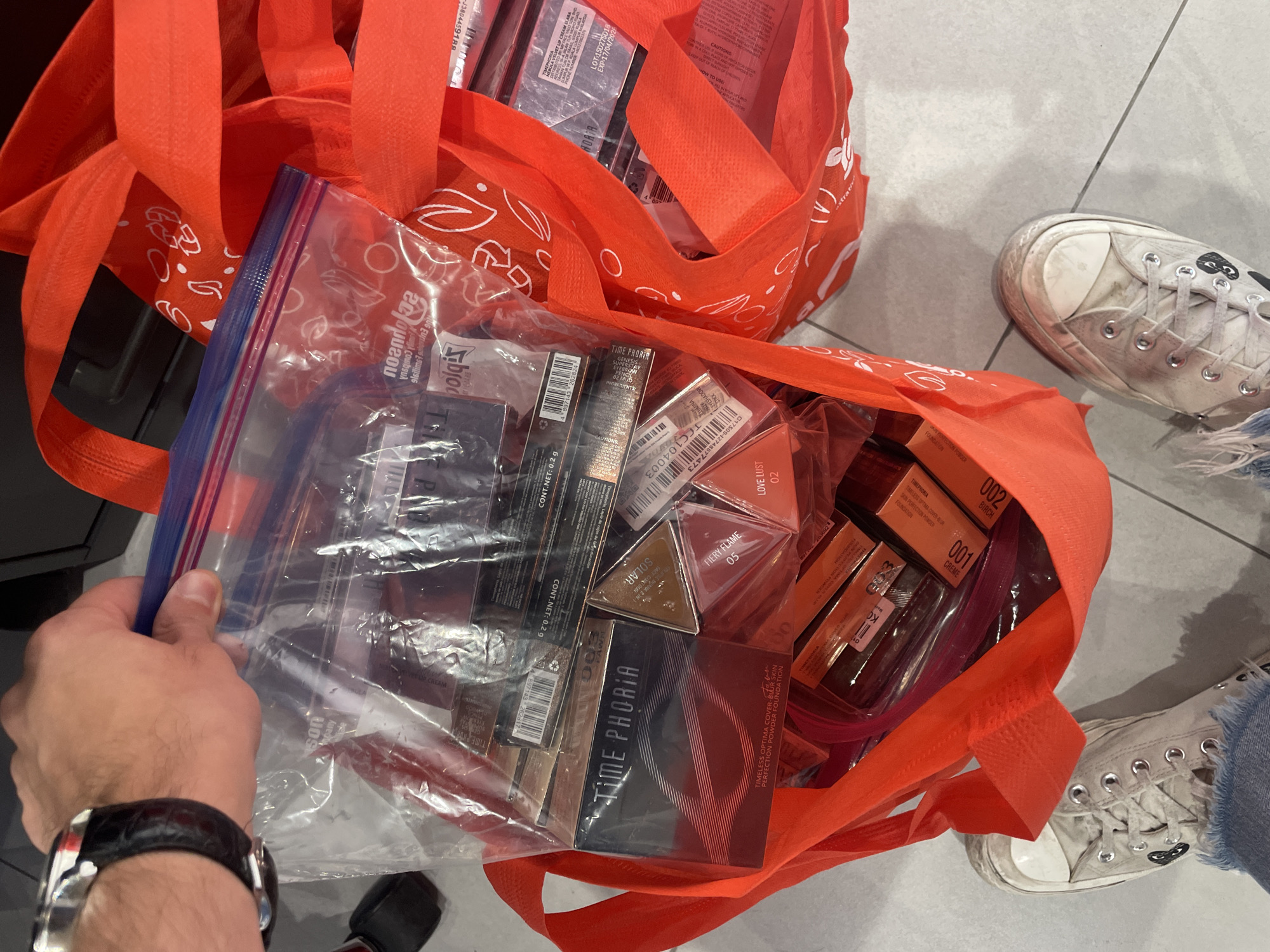 Here is a detailed description of the image:
**Scene:** The image shows a person holding a clear plastic zip-lock bag inside an orange tote bag. Another identical tote bag is visible behind it. The bags appear to be filled with cosmetic products, including various shades of eyeshadow and powder foundations from the brand "Time PHORIA," as indicated by visible product packaging.
**People:** The image includes a view of a person's hand and wrist, adorned with a black leather watch with a silver casing. Another person is visible from the knees down wearing white Converse sneakers and jeans.
**Objects:** The primary objects are two orange tote bags filled with cosmetics, a clear zip-lock bag, and the aforementioned watch. There are cosmetic products like eyeshadow and powder foundations. The orange tote bags are decorated with a bandana pattern.
**Environment:** The setting appears to be indoors, with a tiled floor visible. The floor tiles seem to be light gray. A black object is visible in the lower-left background.
**Location Inference:** While not explicitly stated, based on the context and details, the scene is likely located in Mexico City, Mexico, given the brand "Time PHORIA" specifically references Kuala Lumpur, Malaysia, the cosmetics are likely being sold or distributed in a retail location.