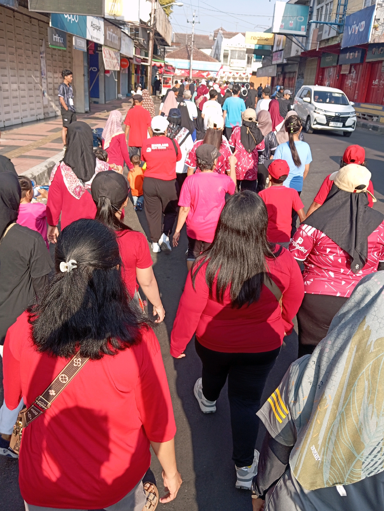 In Magelang, Indonesia, a crowd of people are walking down a street lined with shops. The people are dressed in casual clothing, with many wearing red shirts. Some are carrying bags, and others are holding hands. The street is paved, and there are trees and buildings on either side. The sky is blue, and the sun is shining. A white car is driving down the street in the background. The overall scene appears to be a busy street in a city, possibly during a festival or event, given the large number of people and the festive attire.