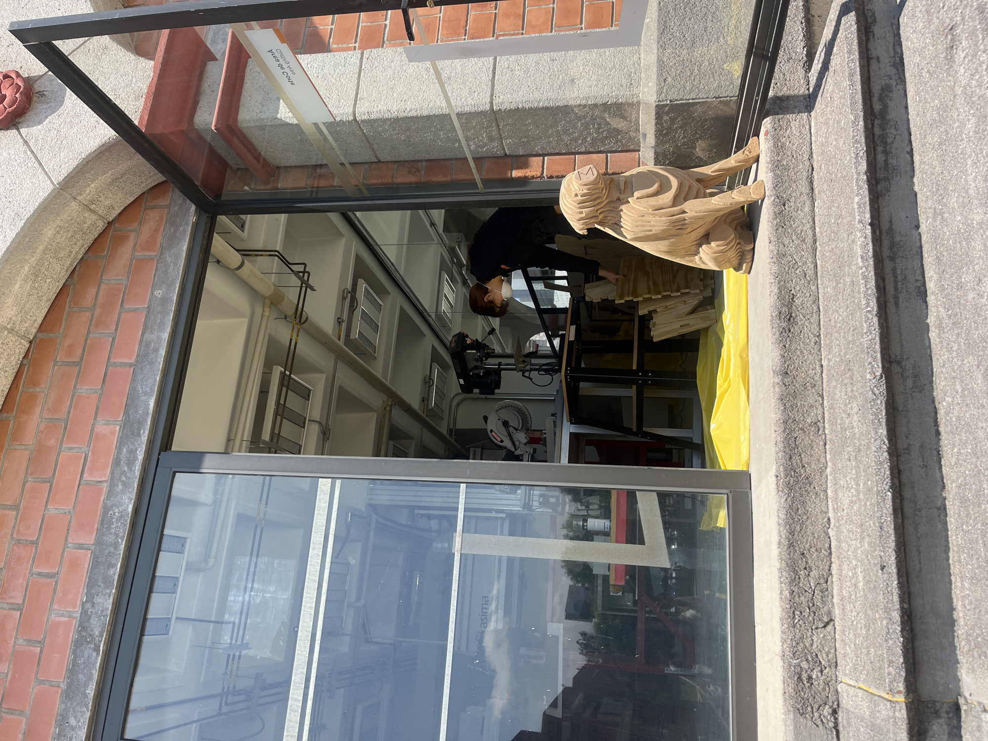 The image shows a street view from the city of San Andrés Cholula, Mexico. It appears to be the exterior of a workshop or studio, seen through a large glass window. Inside, a person wearing a black shirt, mask and safety glasses, is working on a wooden sculpture composed of layered pieces, supported by a black metal frame. There's a woodworking saw and other tools visible in the background. On the exterior windowsill, a finished wooden sculpture is displayed. The architecture of the building includes brick and grey stone elements. Reflections in the window show trees and possibly other buildings outside.