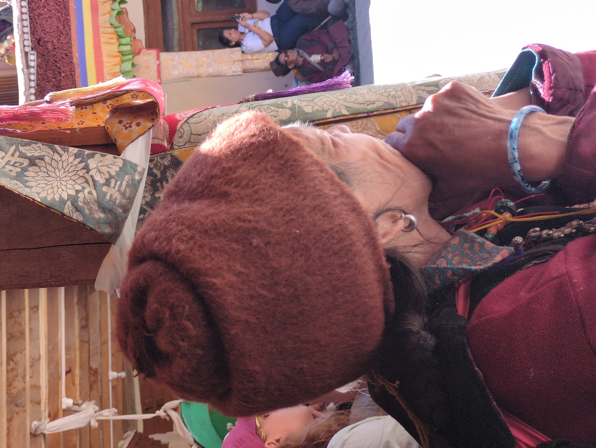 The image shows an outdoor scene, possibly in Pibiting, India, with a focus on an elderly woman in the foreground. She is wearing a large, fuzzy, reddish-brown hat and a maroon jacket with a patterned collar. Her right hand is raised to her face, with a light blue, patterned bracelet on her wrist. To her left, partially visible, is another person wearing a pink hat and a white top. In the background, a woman in a white shirt is holding a phone, and two men are seated on the ground. One man is wearing a maroon robe and a traditional hat, and the other is in similar attire. The environment appears to be a courtyard or outdoor gathering area, with decorative fabrics and architectural elements visible.