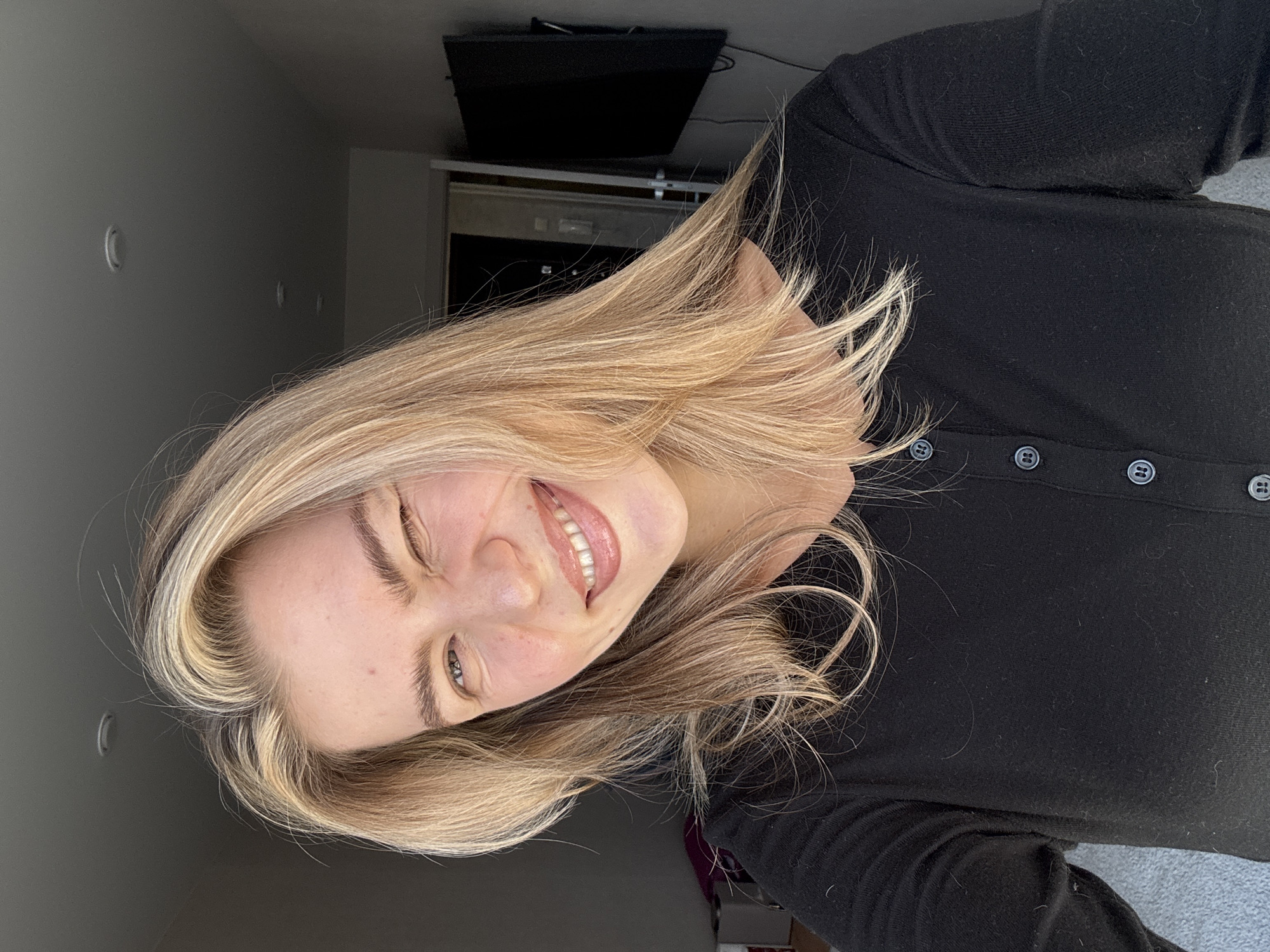 A young woman with blonde hair is depicted in a close-up, head-and-shoulders portrait. She is smiling broadly, showing her teeth, and appears to be winking or squinting her left eye. Her head is tilted, giving the impression of a playful or candid moment. Her hair is a mix of light and dark blonde highlights, styled loosely around her face and shoulders. She is wearing a black, long-sleeved top with several circular, black buttons with silver-toned rims visible down the front.
The background consists primarily of a plain light grey wall. Along this wall, several small, crescent-shaped or circular indentations/protrusions are visible, receding into the distance. In the upper right portion of the background, a dark, rectangular object, possibly a television screen or speaker, is mounted. The lighting is bright, illuminating the woman's face and hair, suggesting a daytime setting. Her expression conveys happiness and cheerfulness. No text is visible in the image.