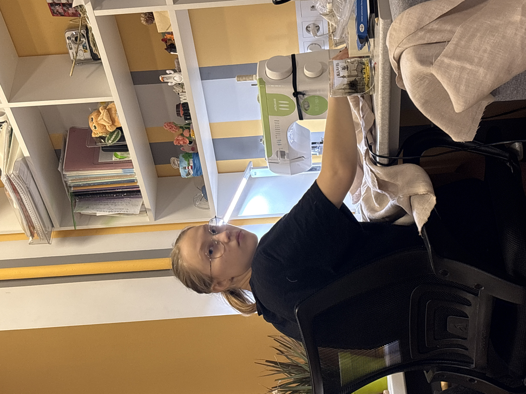The image shows a young woman with blonde hair, wearing glasses and a black shirt, sitting in an office chair. She is facing the camera and appears to be operating a sewing machine, possibly a Jaguar brand, which is positioned on a table. Next to the sewing machine is a small, rectangular object with Russian writing on it, possibly a keepsake or decoration. The table is covered with beige linen fabric. Behind the table is a shelving unit with various items including books, decorative objects, and what appears to be a digital camera. The room has a yellow and white color scheme. The location of the photo may be Perm, Russia as indicated by the text on the object near the sewing machine.