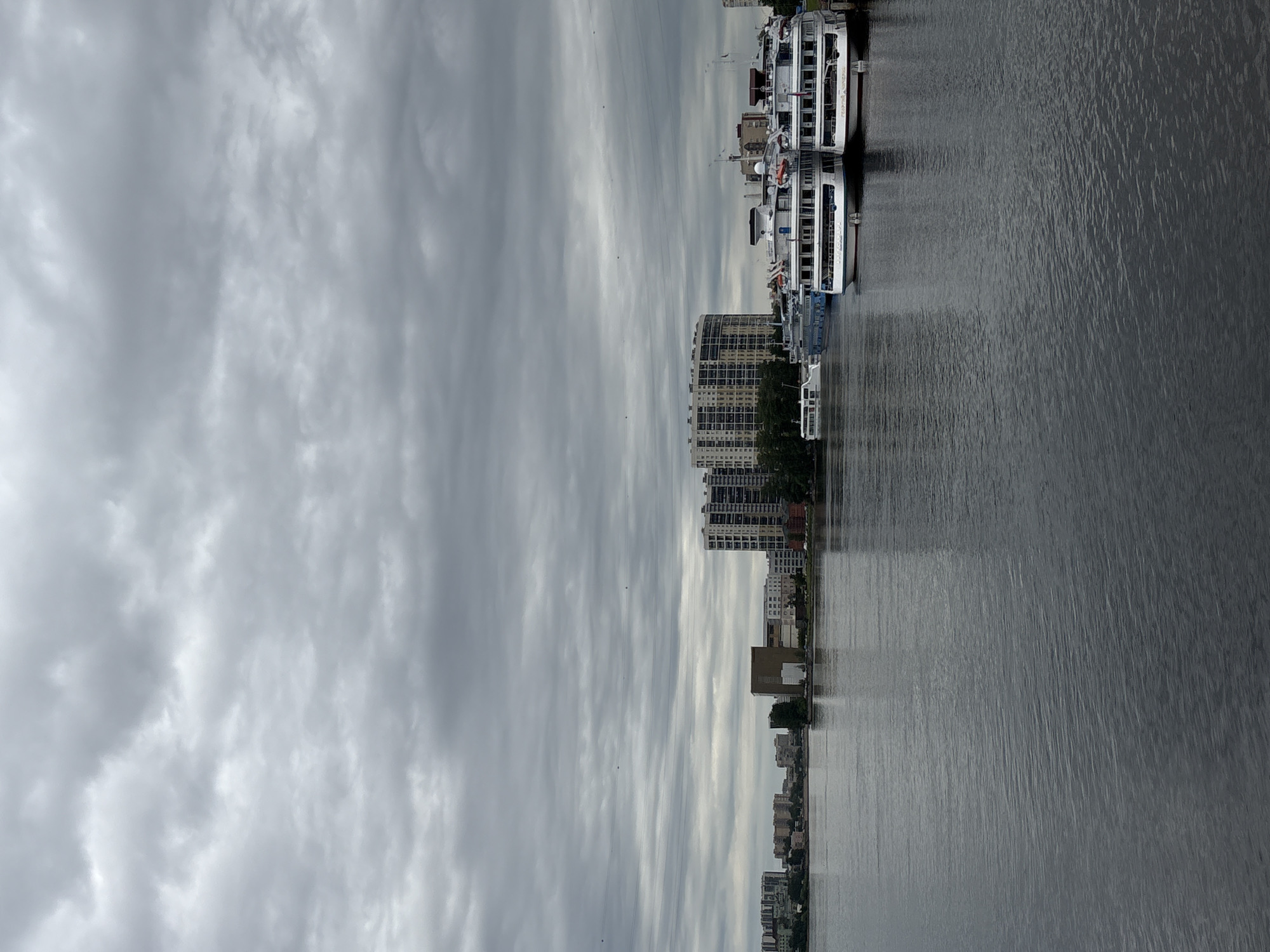 The image depicts a cloudy day over the Neva River in Shushary, Russia. Several large, white passenger ships are docked along the right bank. In the background, a city skyline is visible, with various residential and commercial buildings. The river itself appears calm, with subtle ripples on its surface. The overall atmosphere is somber due to the overcast sky.