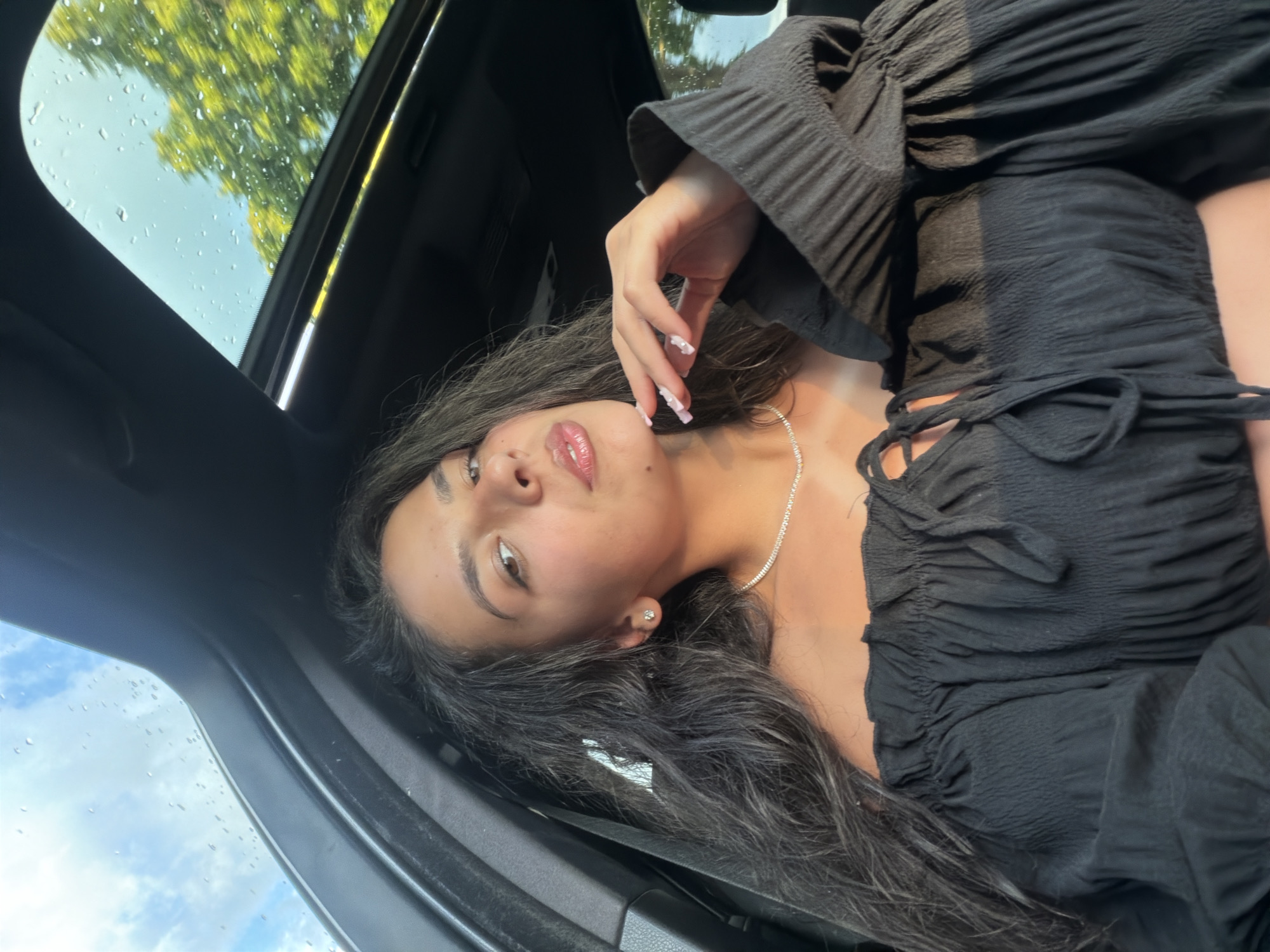 A young woman is captured in a selfie from inside a car, with the sunroof open and rain visible on the glass, suggesting a recent shower. She is wearing an off-the-shoulder black top and a diamond necklace. Her hand is resting on her chin, and she is looking towards the camera with a subtle expression. The background shows greenery and a blue sky with clouds. There is no indication of the city of Veliko Tarnovo, Bulgaria in the image or its description.