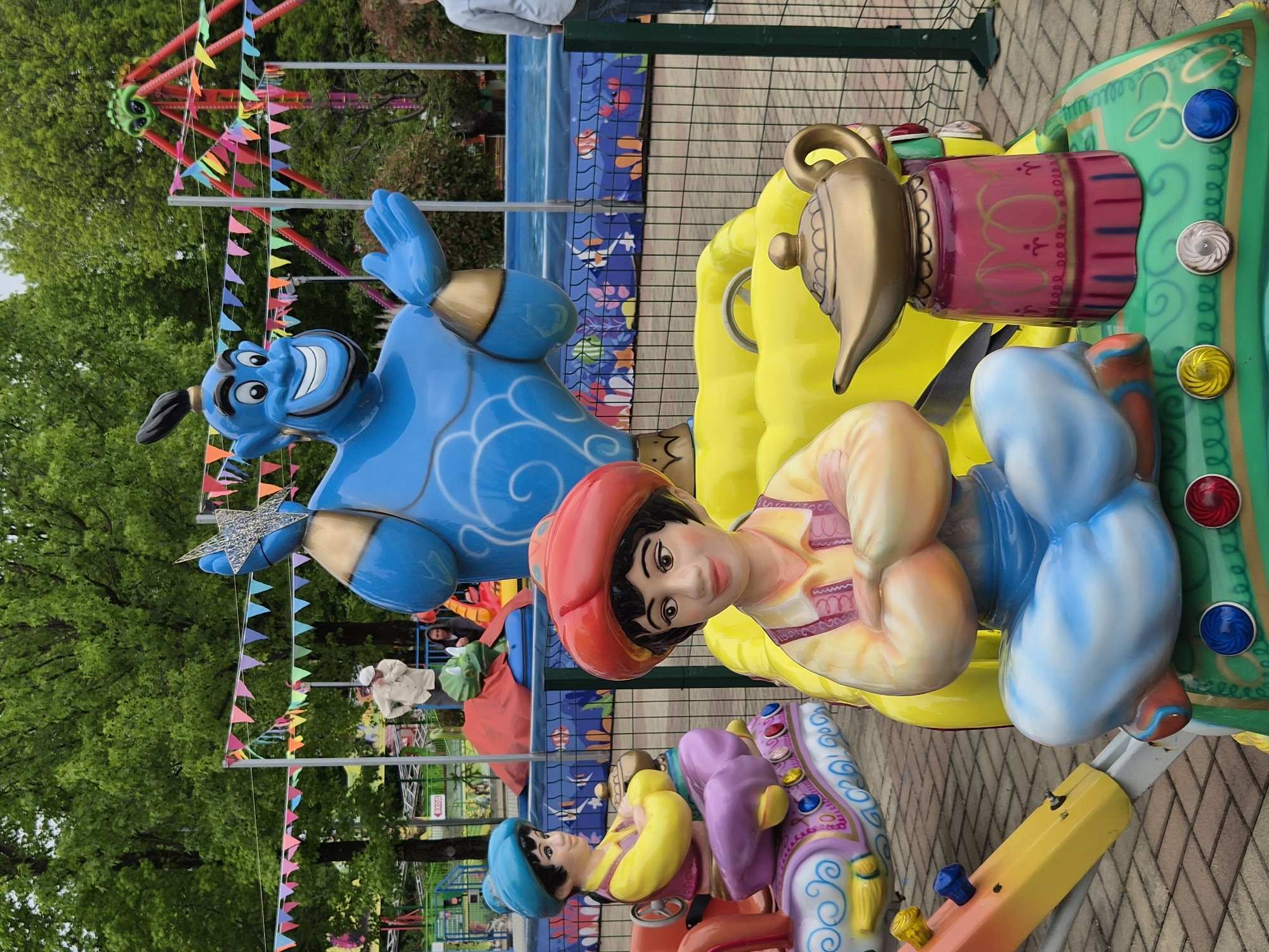 The image depicts a carousel with characters and items from the Disney movie "Aladdin". In the background are trees, a fence, and a ride with multi-colored flags. The foreground contains the Aladdin-themed carousel with depictions of the Genie, Aladdin, and a magic lamp. The location is likely an amusement park or recreational area in Sochi, Russia.