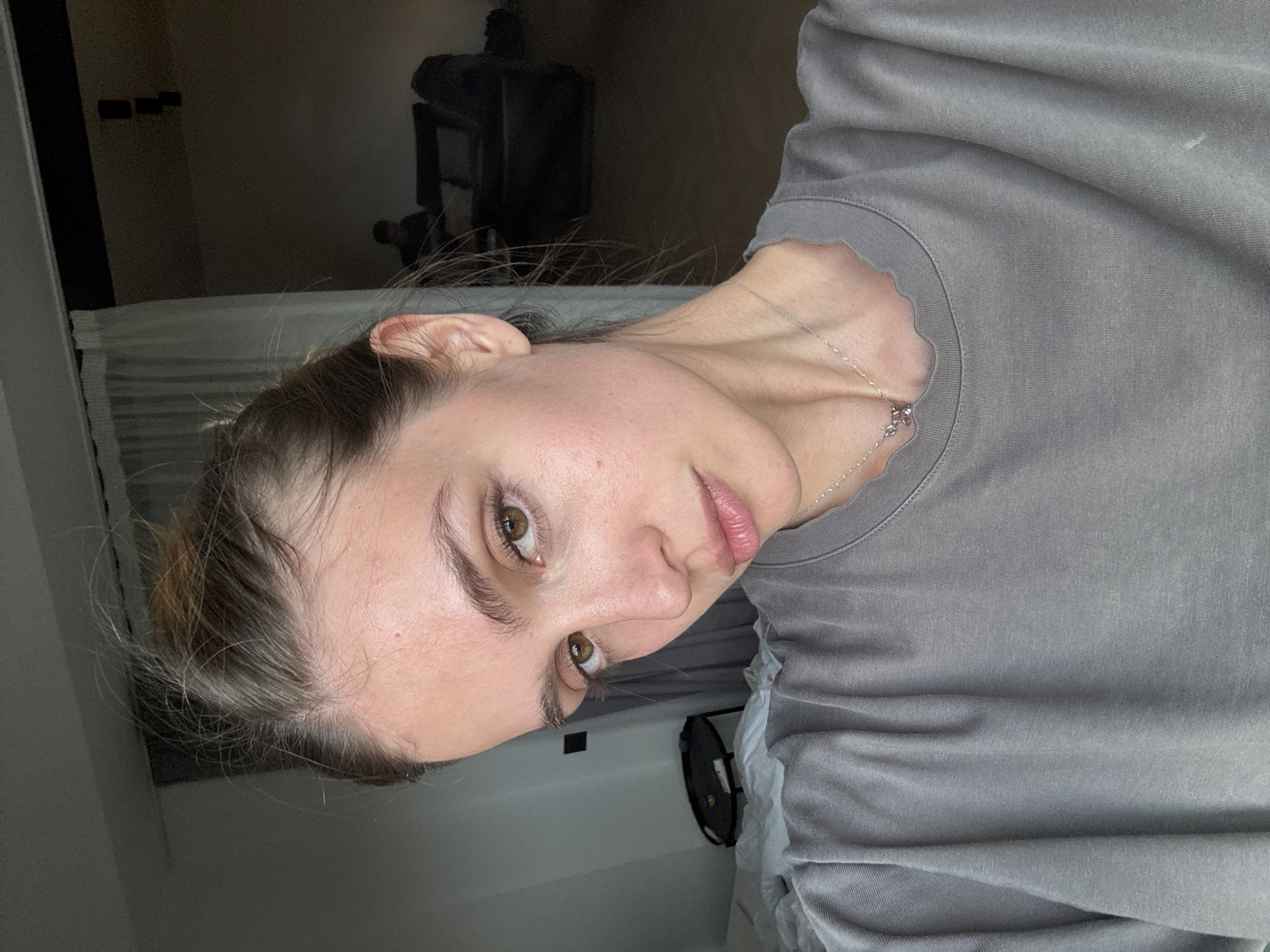 A woman with brown hair pulled back into a bun and light skin looks directly at the camera with brown eyes. She wears a gray shirt and a silver necklace. The background is a room with a bed and a piece of furniture. The exact location in Yerevan, Armenia cannot be determined from the image.