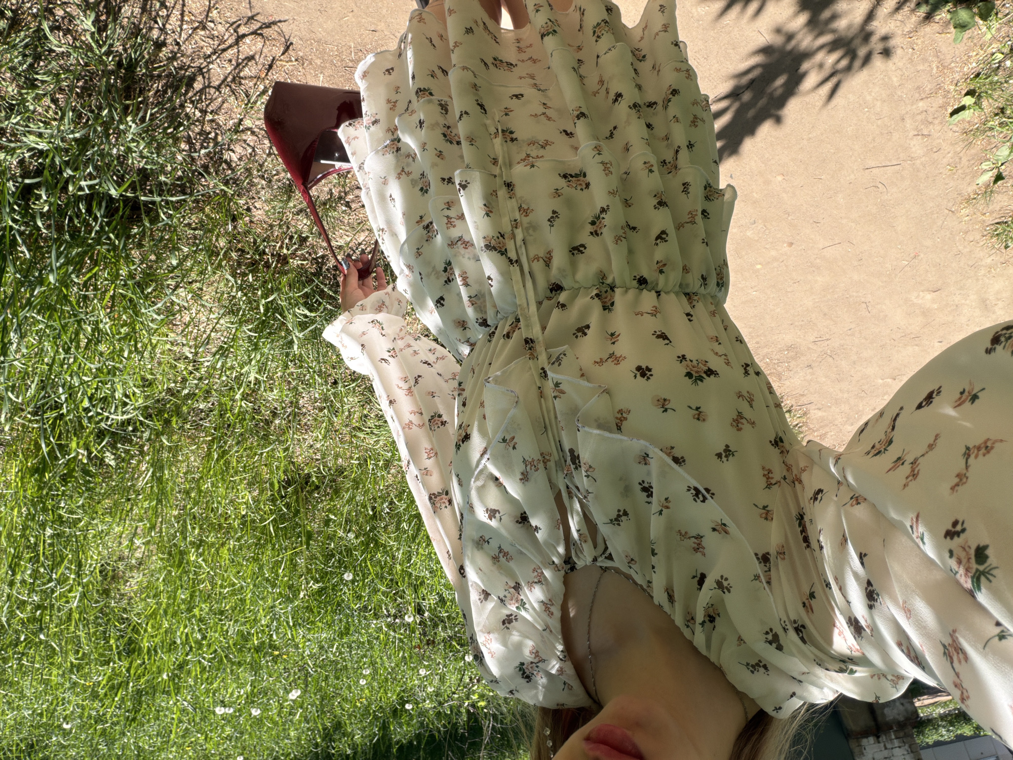 A woman in a white floral dress is holding a burgundy purse while standing on grass in an outdoor environment. Part of a dirt path is visible in the upper right of the frame, with some trees casting shadows on it. The city is unknown.