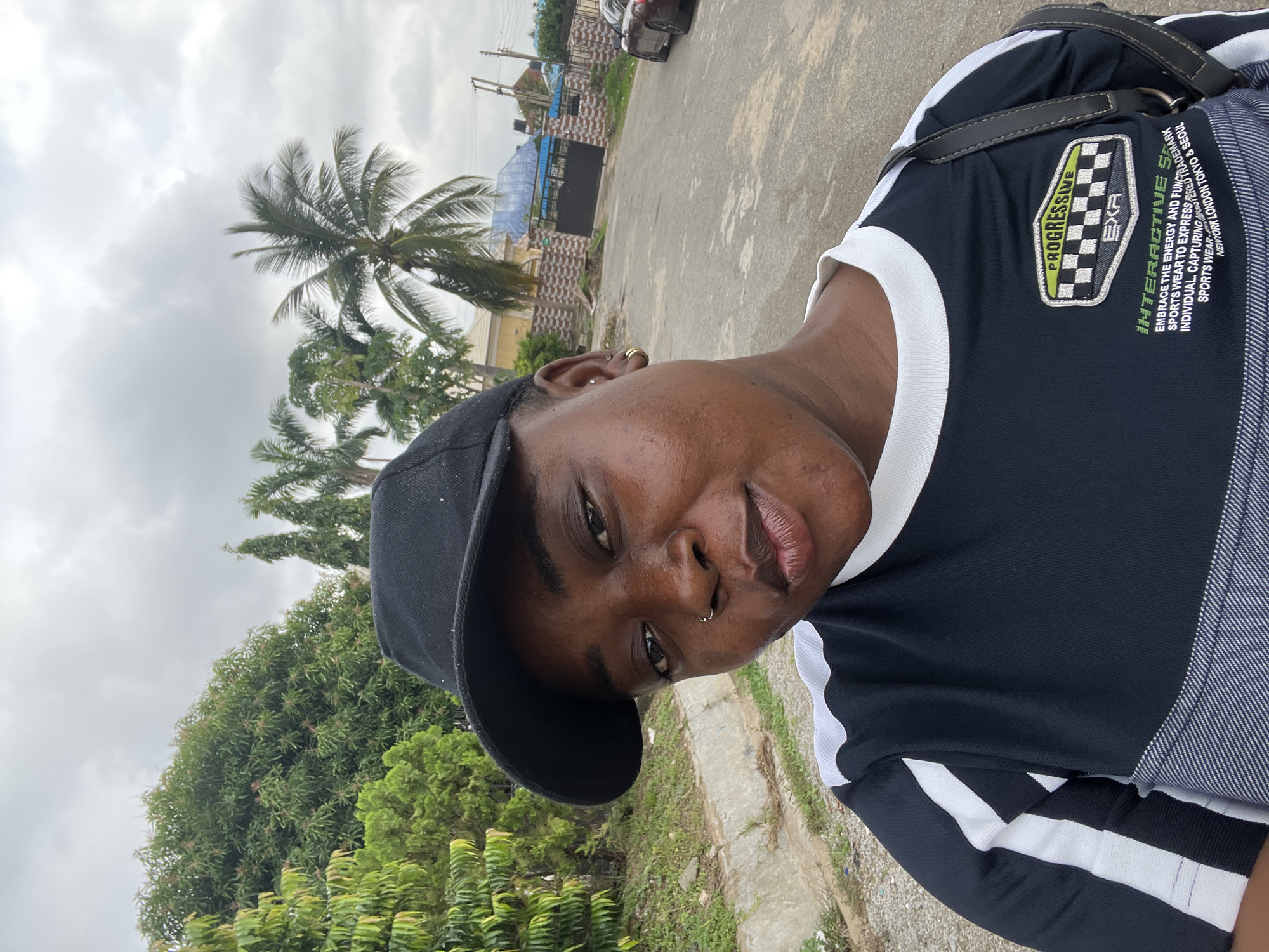 A medium shot shows a person wearing a black baseball cap and a black and white striped t-shirt. The person has dark skin and is looking at the camera. The background is a street with trees and houses. The sky is cloudy. The image is taken outdoors in Abuja, Nigeria.