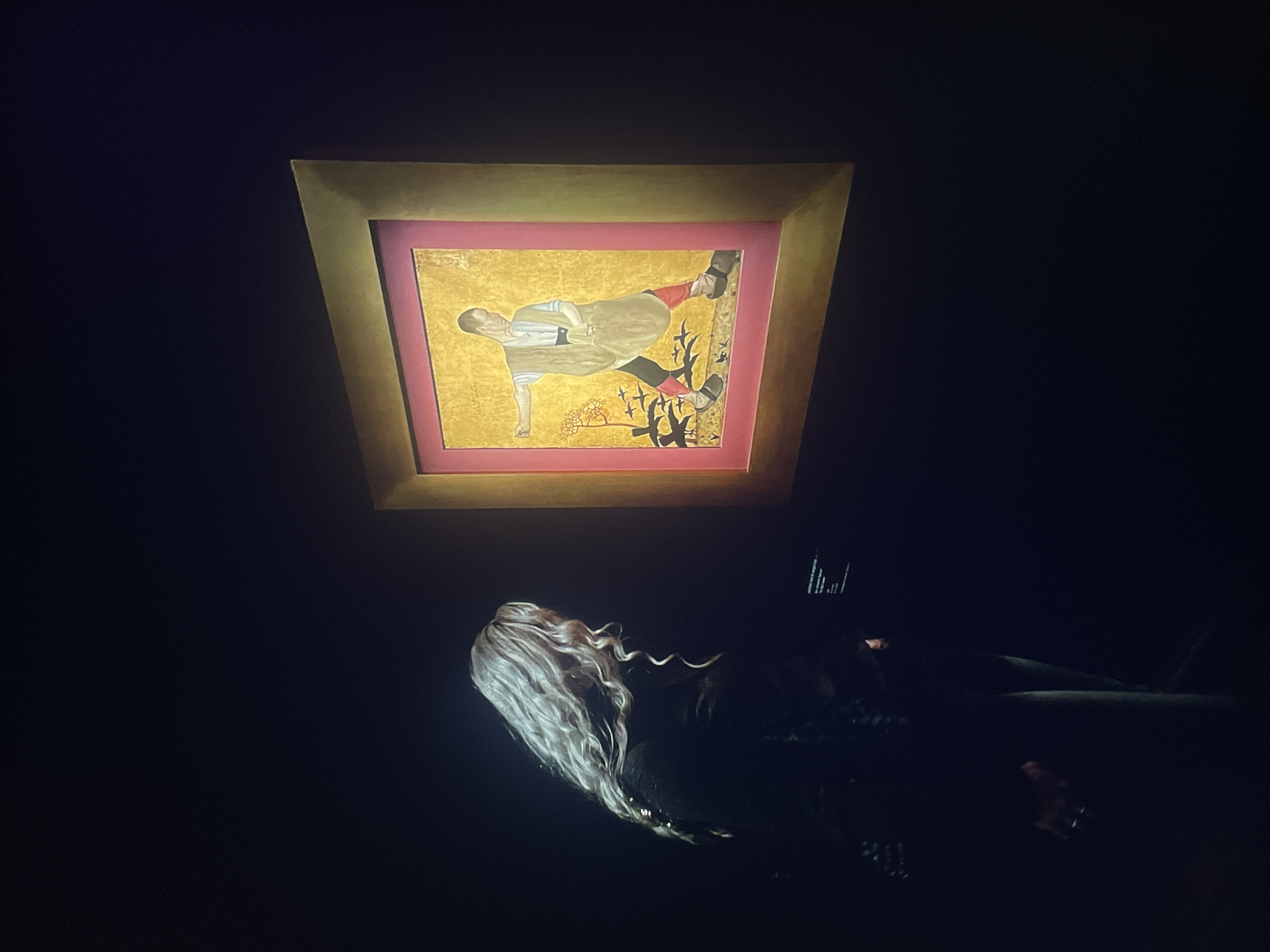 A woman with long, wavy white hair is lying down, looking up at a square-shaped painting hanging above her. The painting has a gold background, a pink border and a gold frame. The painting depicts a man in tan clothing, red stockings, and black shoes, surrounded by a small tree. The lighting is dim. The location is the city of Unknown, Unknown.