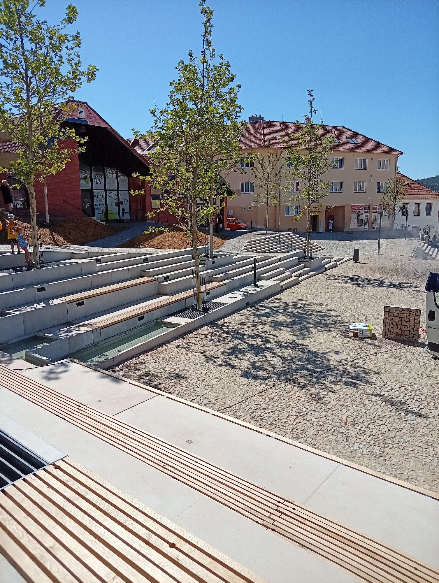 In Tišnov, Czechia, a modern public square is depicted under a clear blue sky. The scene features a multi-tiered seating area constructed from concrete and wood, interspersed with young trees. Water features are integrated into the lower tiers of the seating. The ground of the square is paved with cobblestones. In the background, brick and stucco buildings with red tiled roofs line the square. A red car is parked on the left, and a van is visible on the right. Several people are present, including two children standing on the upper steps of the seating area. The overall impression is one of a newly developed or renovated urban space designed for public gathering and relaxation.