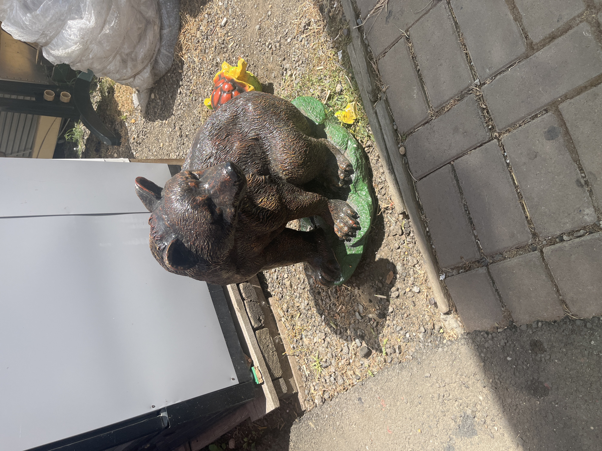 In Plastunovskaya, Russia, a weathered, brown, bear-shaped garden ornament sits on a green base that mimics a leafy surface. The ornament is positioned on a ground covered with small stones and dry grass, adjacent to a grey paved walkway. Behind the bear ornament, a large white plastic bag is visible, partially obscuring a dark green metal structure. The scene is illuminated by bright sunlight, casting shadows on the ground.