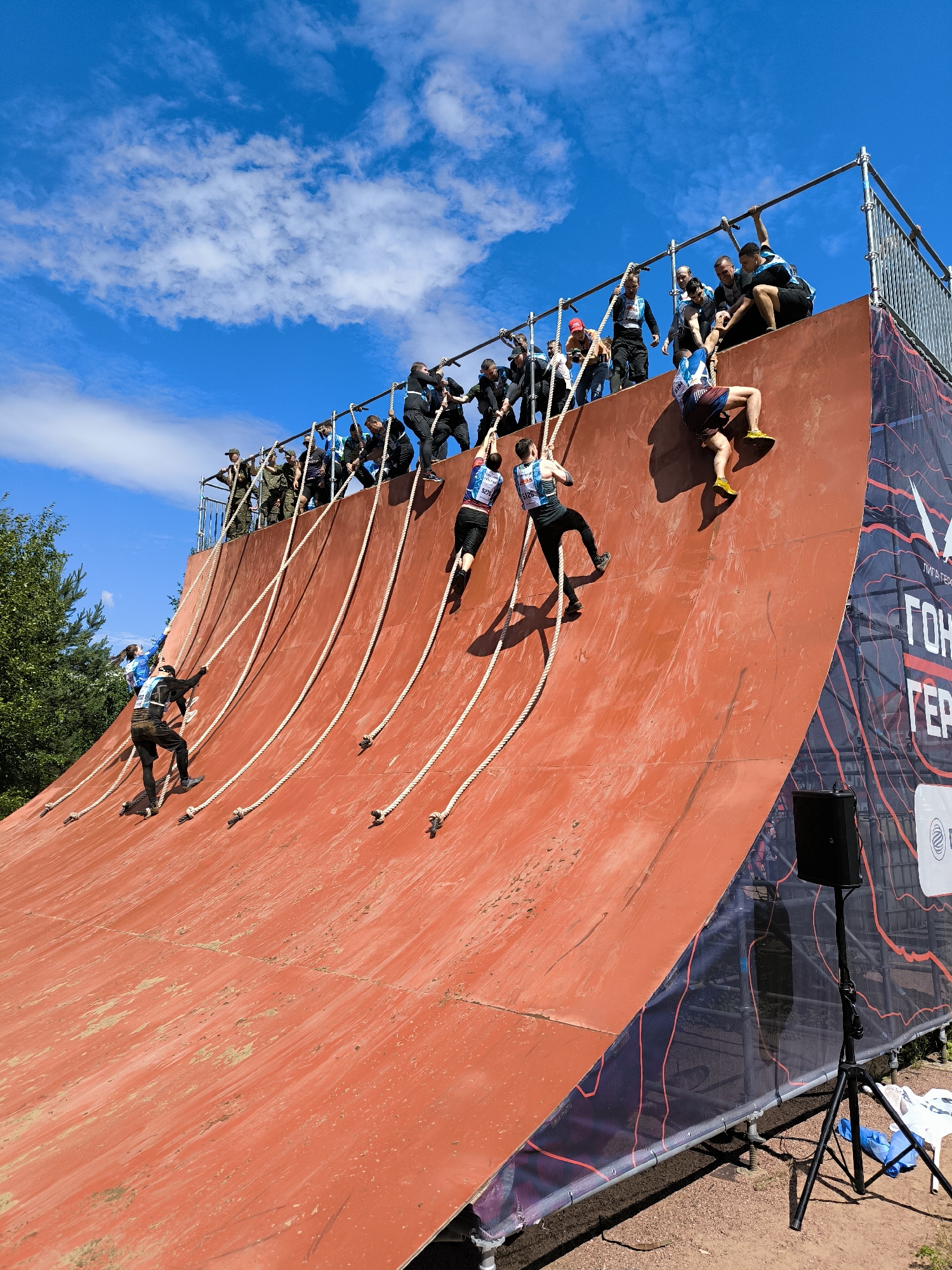 The image depicts an outdoor obstacle course in Sertolovo, Russia. A group of participants are scaling a large, red, curved wall using ropes. Some individuals are near the top of the wall, being assisted by others standing on a metal platform. A few participants are lower down, actively climbing. The environment includes a bright blue sky with scattered clouds, trees in the background, and a dirt ground surface. There is also a speaker on a tripod and some banners with text, one of which reads "Гонка Героев," indicating the event's name, which translates to "Race of Heroes."