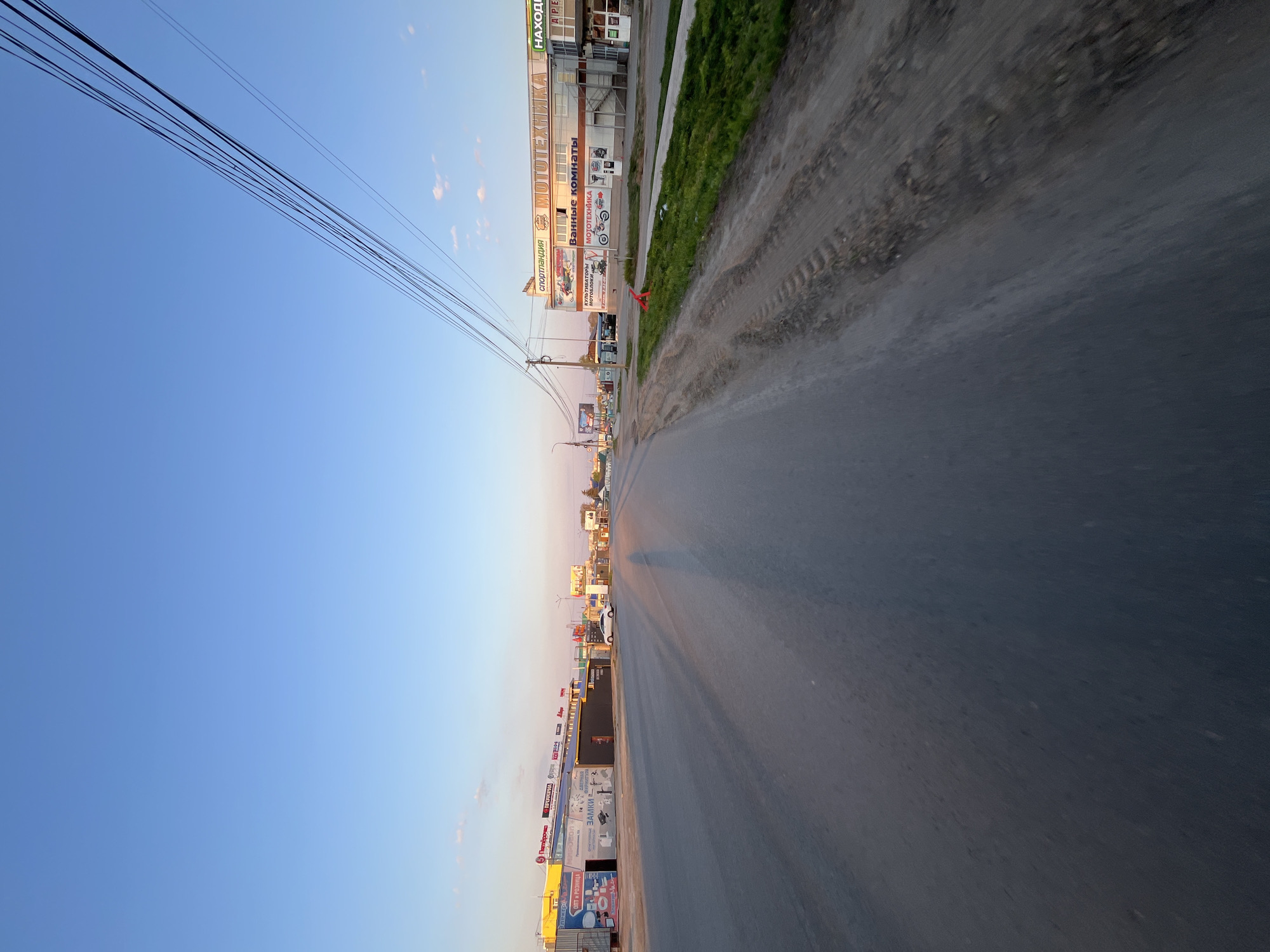 The image shows a street view in the city of Chaikovsky, Russia. The road stretches into the distance, flanked by buildings and greenery on the right, and open sky with power lines on the left. There are visible commercial buildings with signage along the road. The road itself is asphalt, showing signs of wear. The lighting suggests it is either early morning or late evening.