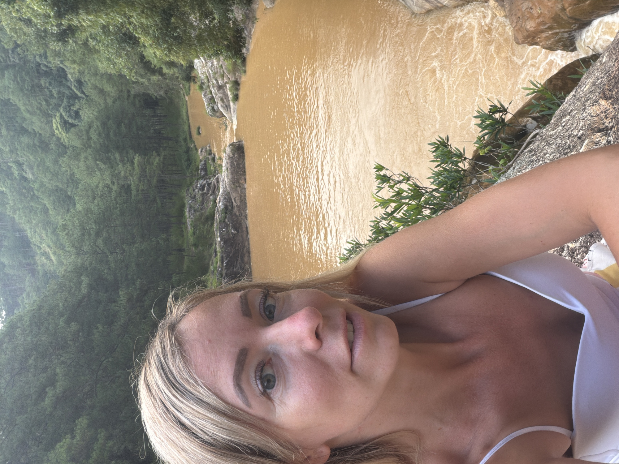 In a selfie taken outdoors, a blonde woman with blue eyes and fair skin is positioned in the foreground, looking directly at the camera. She is wearing a white camisole that exposes her shoulders and décolletage. Her hair is styled in loose waves. Behind her, a murky, brownish-orange river flows through a rocky gorge. The riverbanks are lined with lush green trees and vegetation, with a dense forest extending up the hillsides in the background. The overall setting appears to be a natural landscape, likely in the vicinity of Păng Tiêng 1, Vietnam, given the tropical appearance of the foliage and terrain.