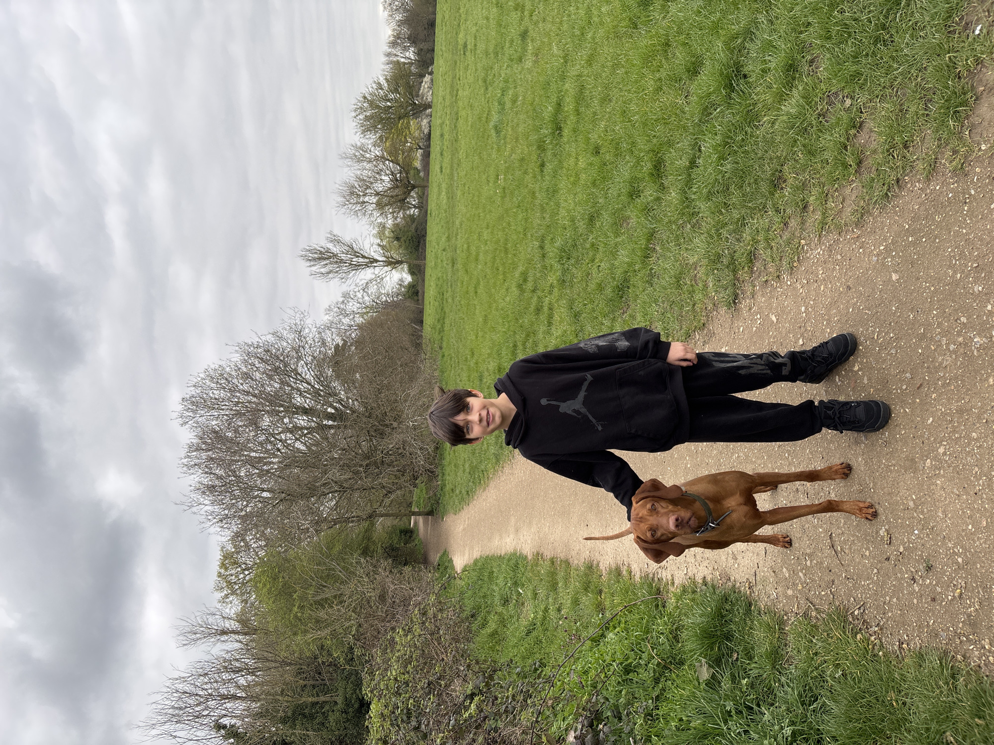 A young person in a black hoodie and black pants stands on a dirt path with a short-haired brown dog. The path is surrounded by green grass and trees. The sky is overcast. The location is Broxbourne, United Kingdom.