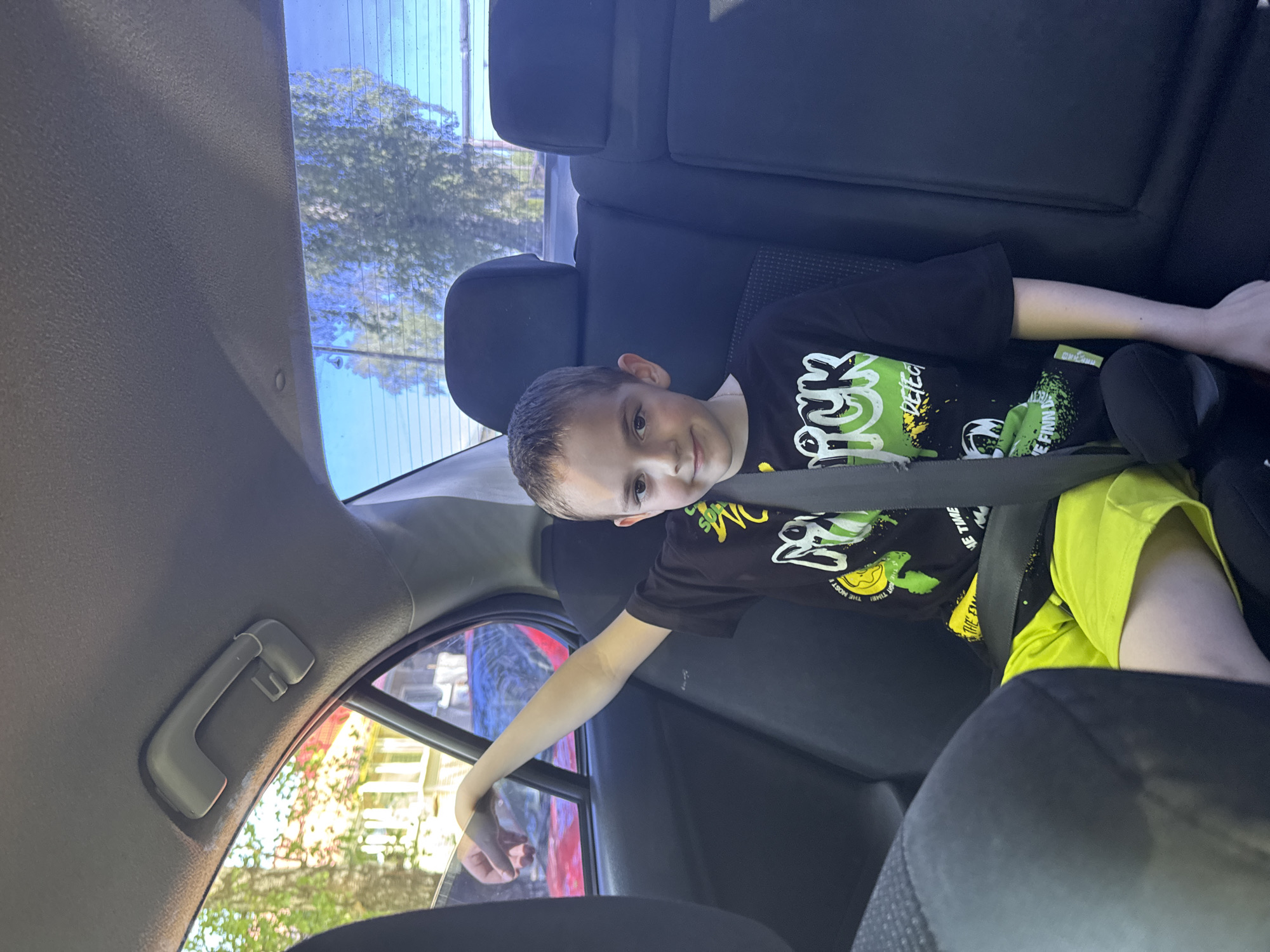 A boy is seated in the back seat of a car, wearing a black t-shirt with a green and white design and neon yellow shorts. He is wearing a seatbelt. His left arm is extended towards the car window. Outside the window, there are trees and a red car. The car is parked on a street, possibly in a residential area. I cannot confirm the location is in Раздолье, Russia.