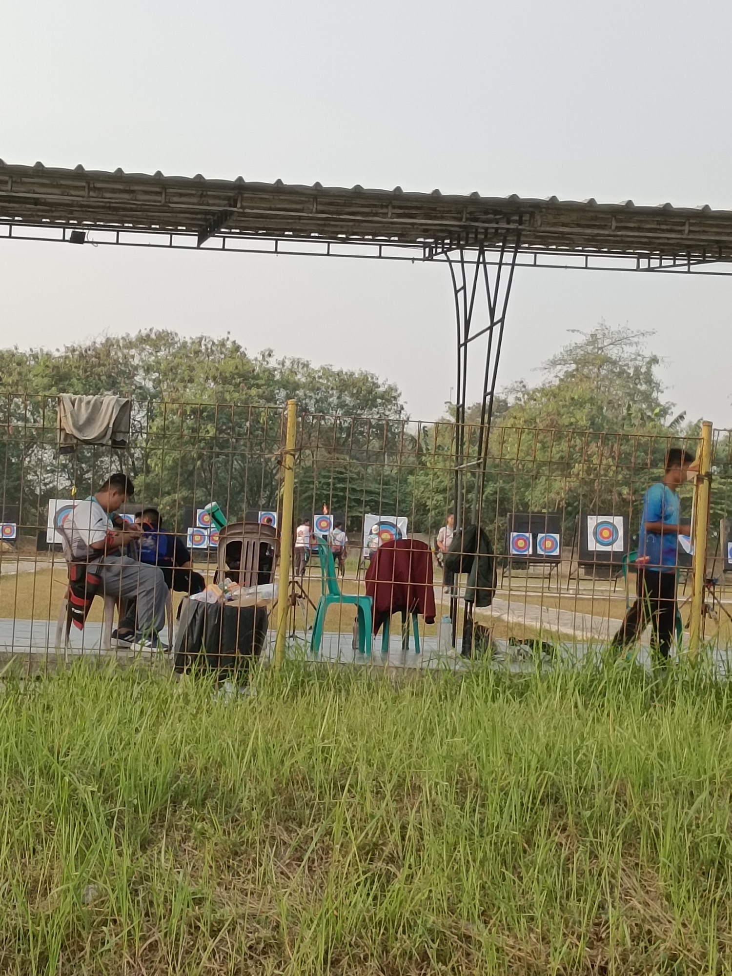 The image depicts an archery range in Sertajaya, Indonesia, on a bright, clear day. The foreground is dominated by a dense, tall growth of green grass. A wire fence runs horizontally across the middle ground, separating the foreground from the archery range. Beyond the fence, several people are visible, engaged in archery-related activities.
On the left side of the frame, two men are seated on chairs. One man, wearing a white t-shirt and grey pants, appears to be adjusting equipment. The second man, in a blue shirt, is partially visible behind him. A towel is draped over the fence near them. Several archery targets, circular with concentric rings of yellow, red, and blue, are set up in the background.
To the right, a young man in a blue t-shirt and black pants is walking away from the camera, holding a piece of paper. Another person in a light blue shirt is visible further in the background. Archery targets are also present in this section of the range. The background is filled with lush green trees. A metal roof structure with a corrugated surface is supported by vertical metal poles, providing shade over part of the archery range. Two green plastic chairs are placed near the fence, and a dark trash bin is situated behind them. The overall impression is a recreational or training facility for archery.