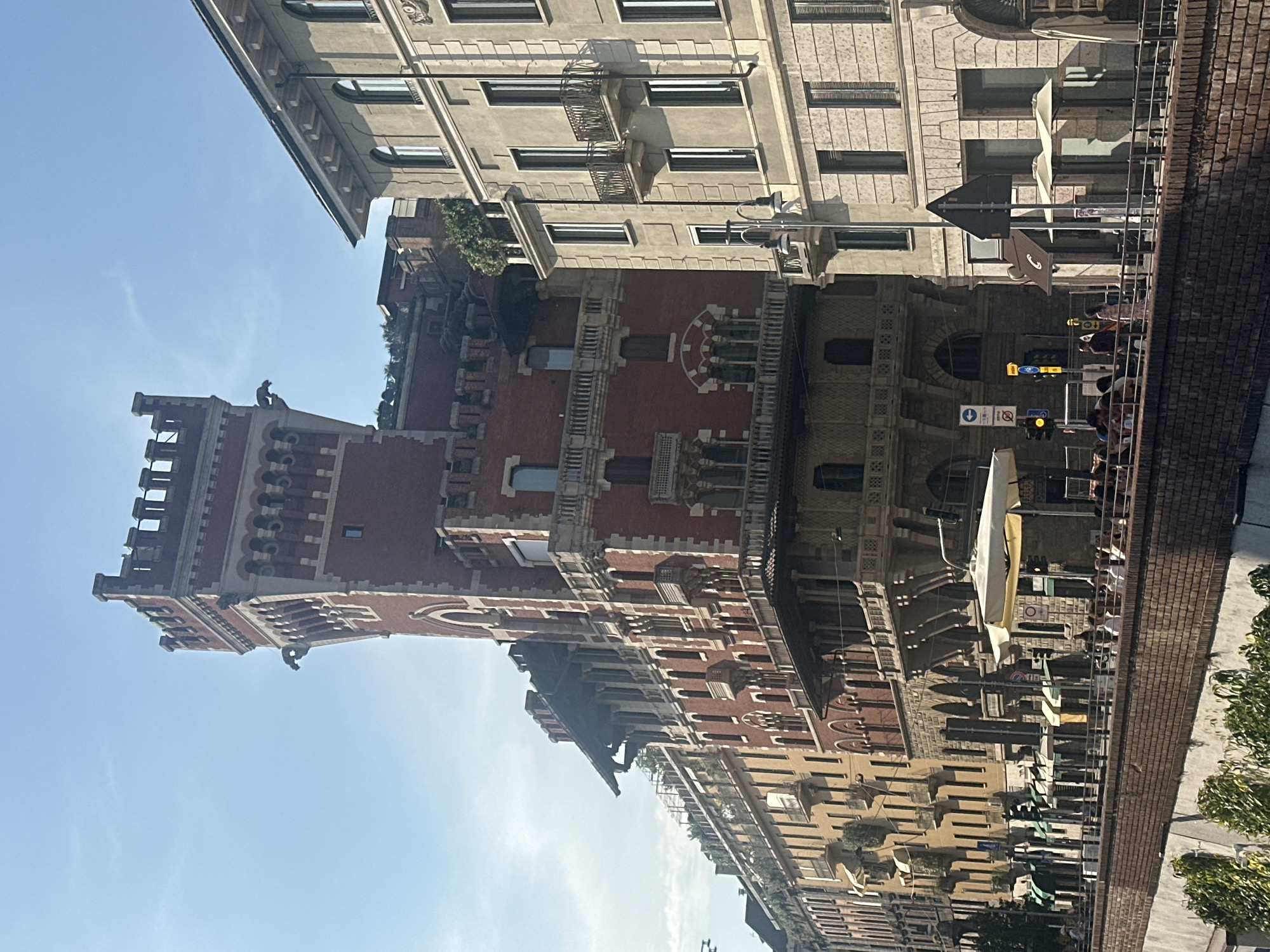 The image shows a street scene in Milan, Italy, with a focus on several buildings and a canal or river. The most prominent structure is a tall, red brick building with a castle-like tower, featuring decorative battlements and what appear to be gargoyle-like figures. This building is flanked by other multi-story structures with balconies and numerous windows.
In the foreground, a brick wall borders a body of water. A promenade or sidewalk runs alongside the water, with a metal railing. Several people are visible on this promenade, some walking and others gathered near tables. There are umbrellas providing shade, suggesting an outdoor seating area for a cafe or restaurant. Traffic lights and street signs are visible, indicating an urban environment. The sky is clear and blue, with a hint of sunlight casting shadows on the buildings. The overall impression is a sunny day in a bustling city.